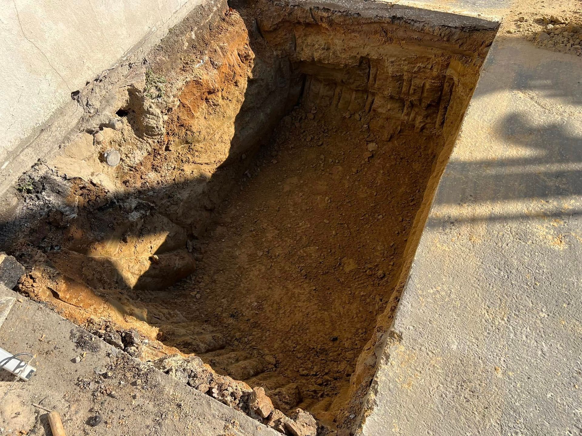Large rectangular hole dug beside a sidewalk, with exposed soil and broken pavement edges