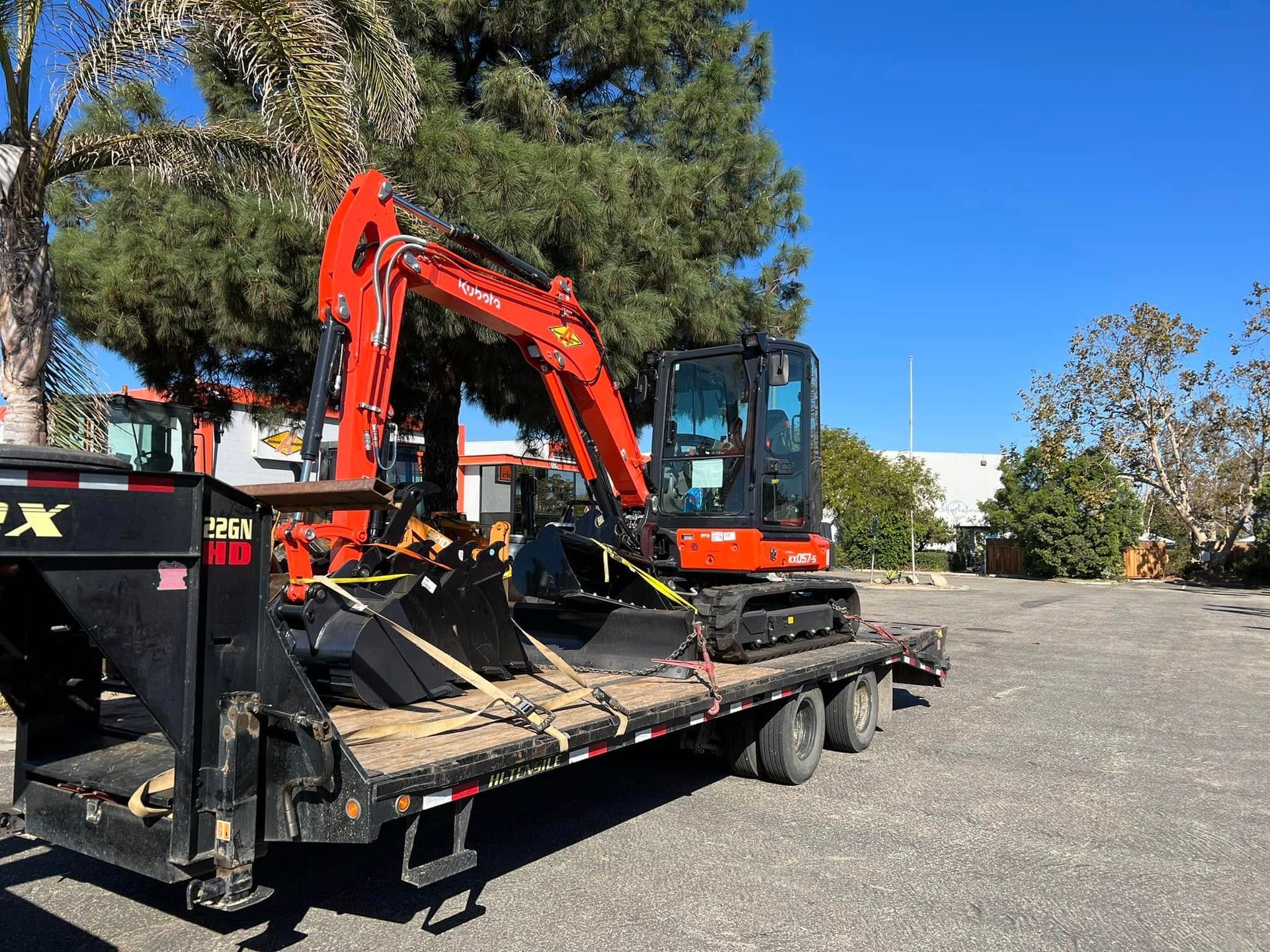 Orange excavator on a flatbed trailer in a gravel lot under a blue sky.