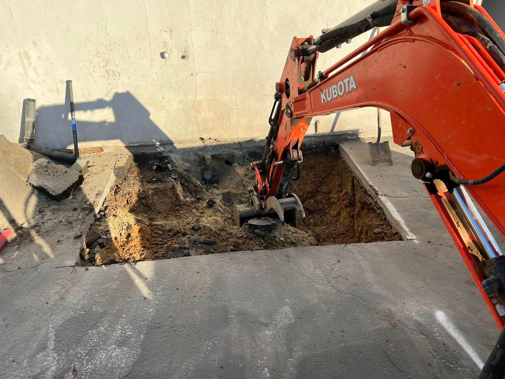 Orange excavator digging a trench beside a concrete wall and paved surface.