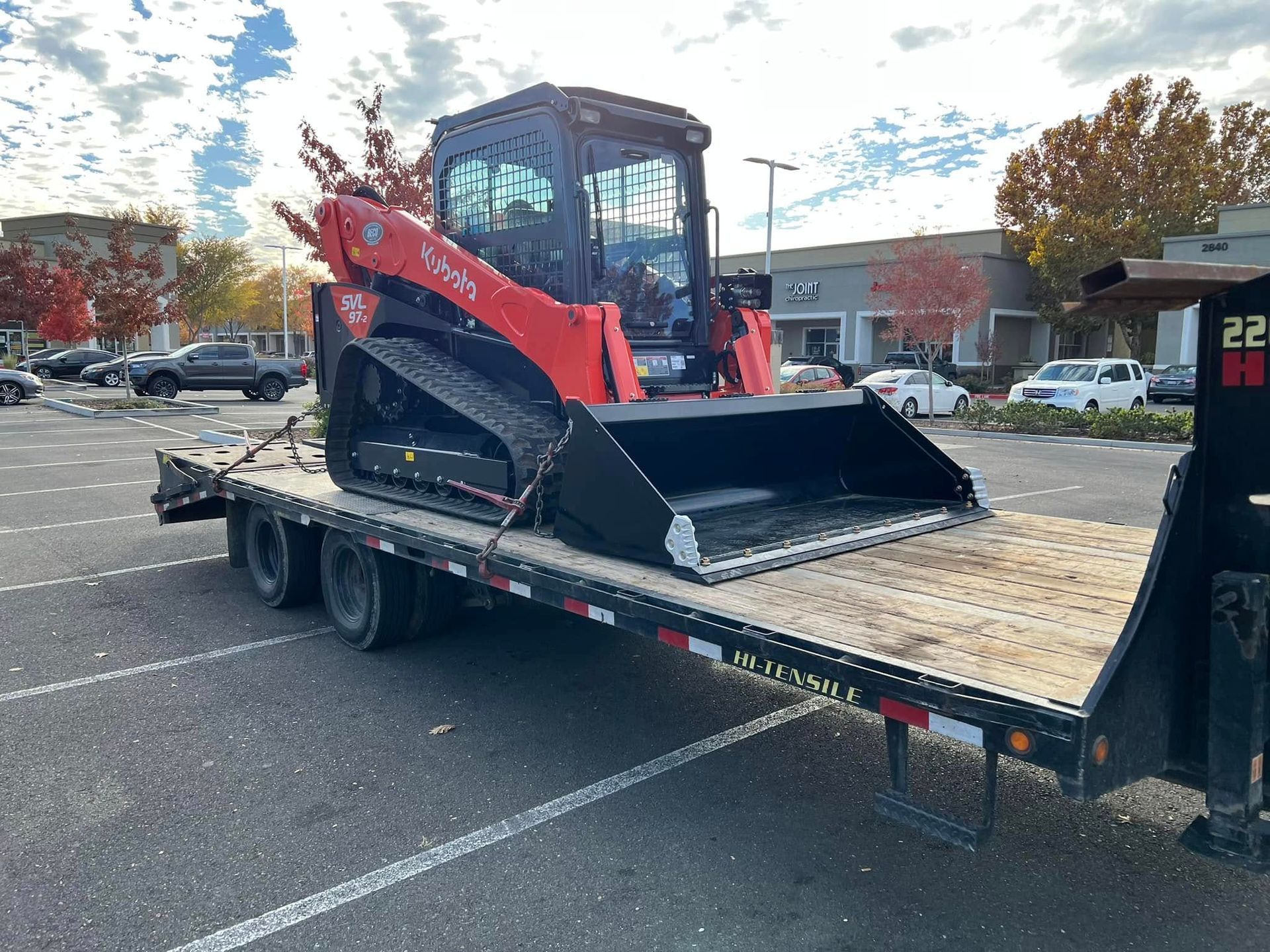 Red skid-steer loader strapped on a flatbed trailer in a parking lot.