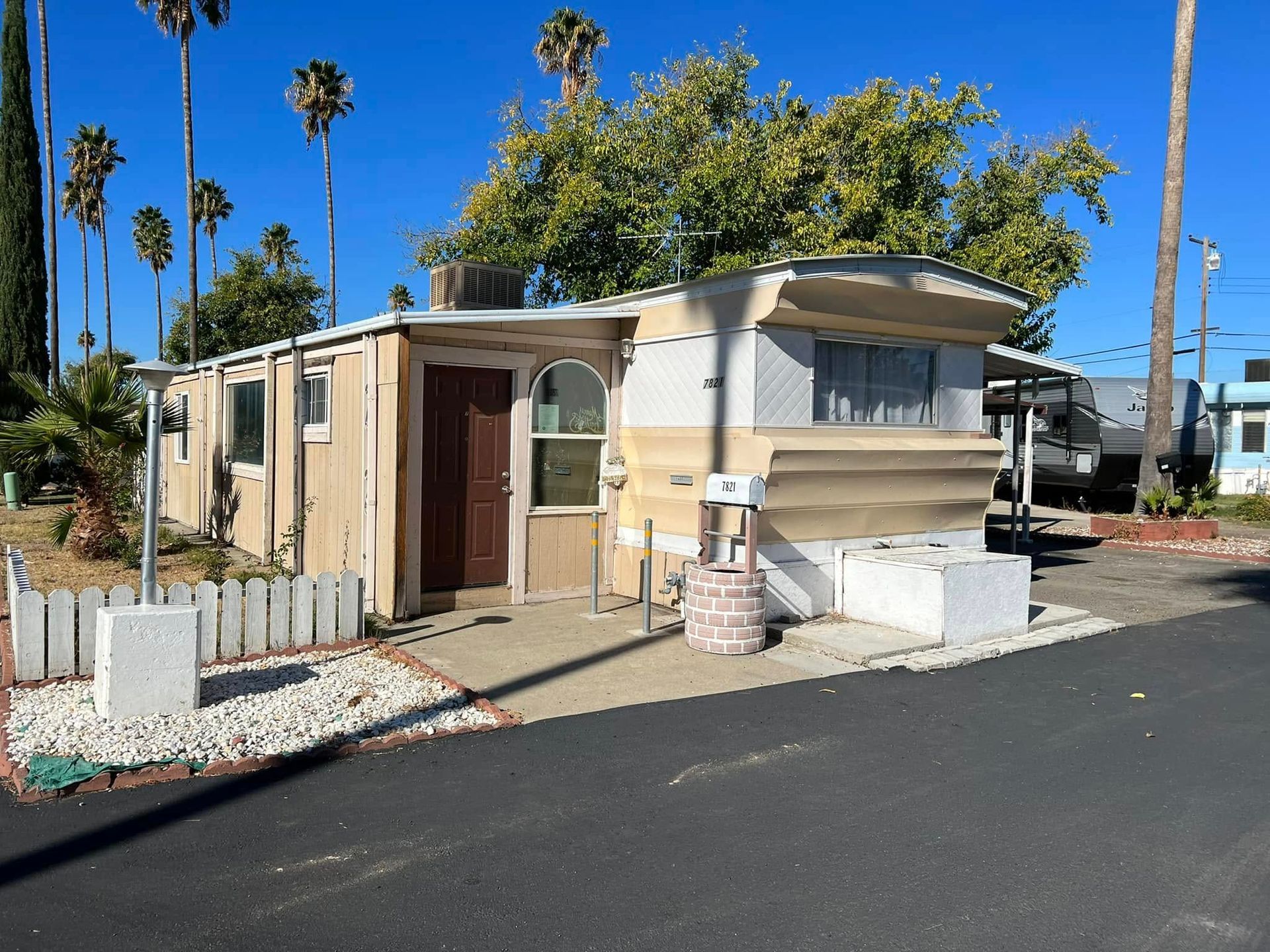 Small beige mobile home with awning and white trim on a sunny street, framed by palm trees.