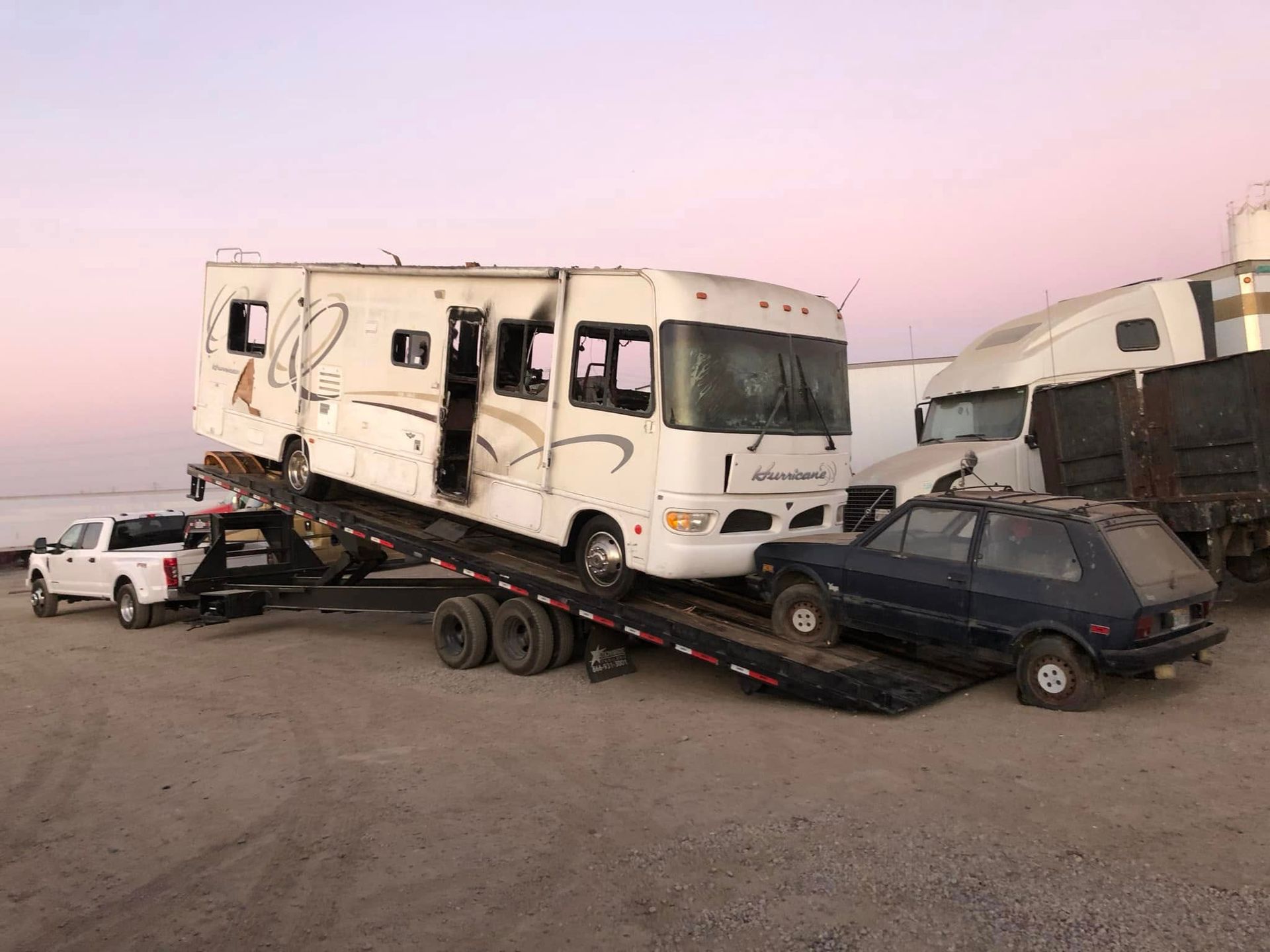 RV and car crash scene with wrecked vehicles on a flatbed trailer in a dusty lot at sunset