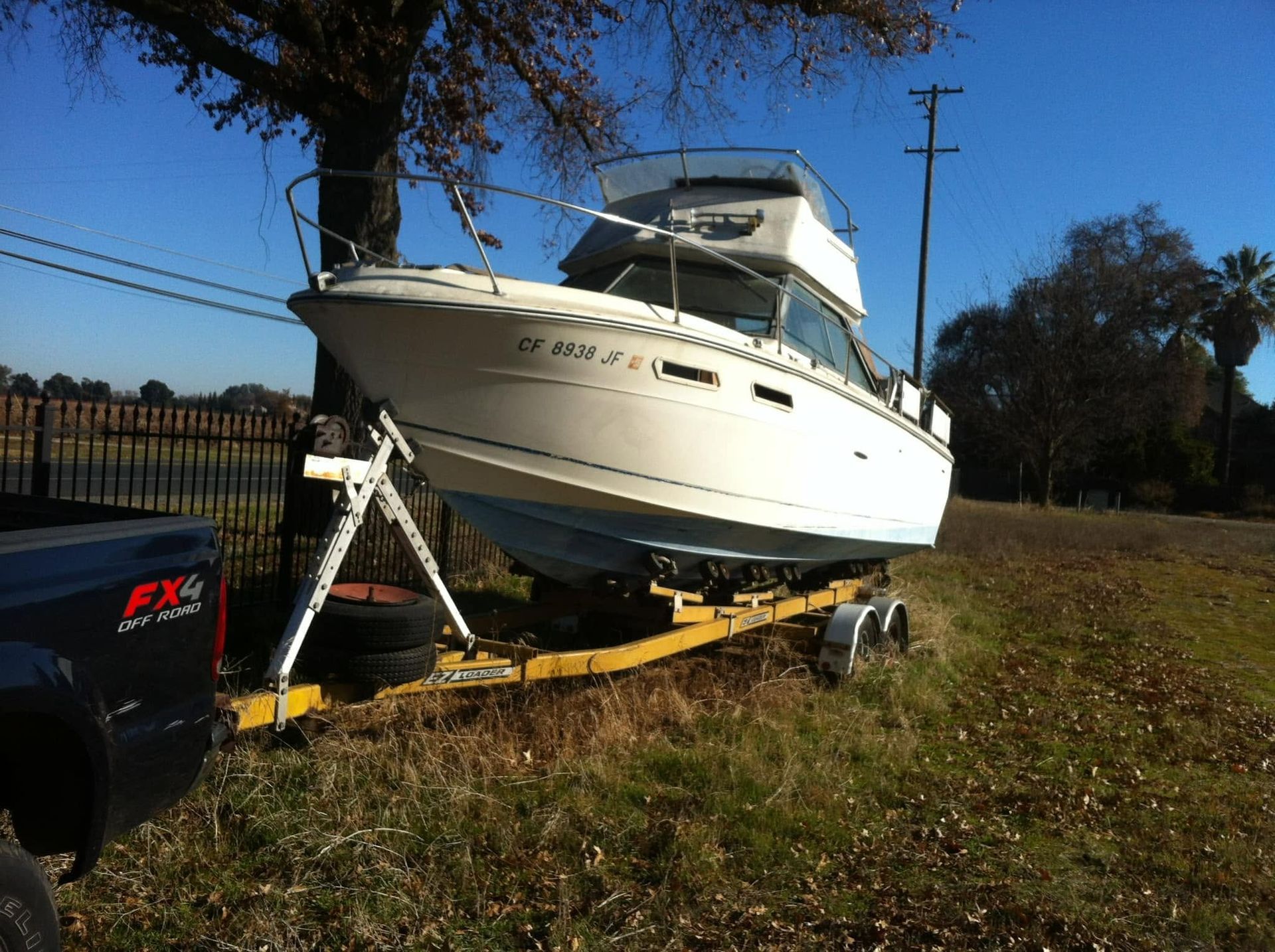 White yacht on a trailer in a grassy yard beside a black pickup truck on a sunny day