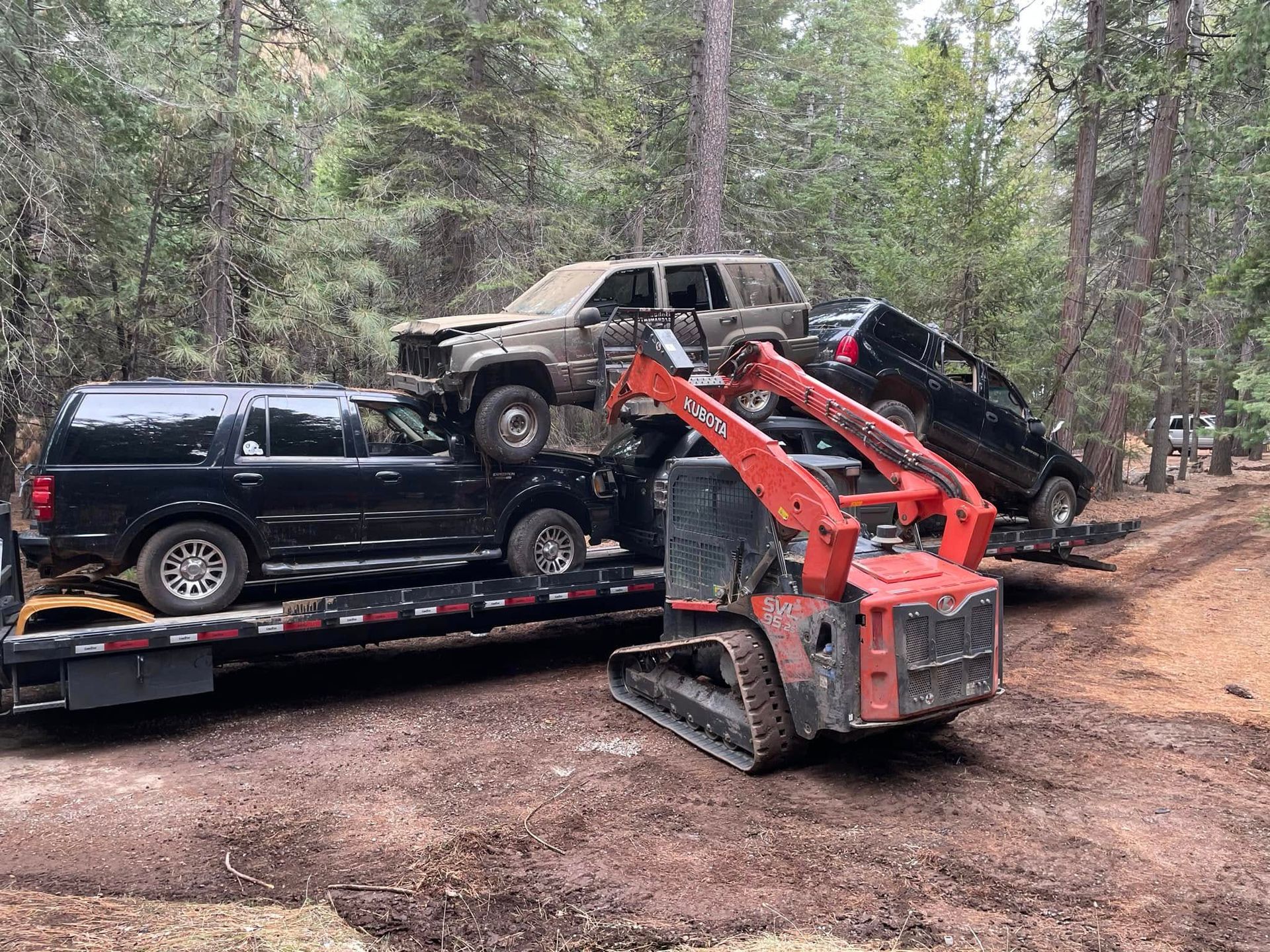 Orange mini skid steer recovering black SUV and overturned vehicles in a wooded off-road area