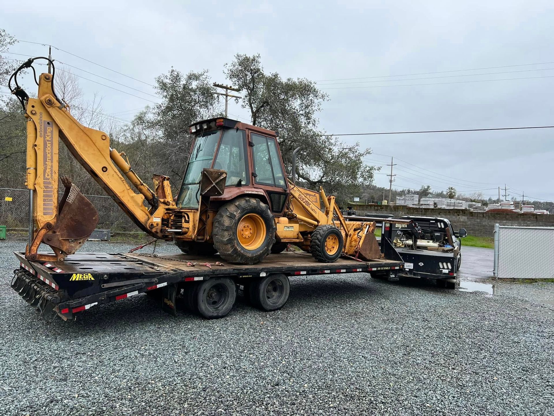 Yellow backhoe on a flatbed trailer in a gravel lot under cloudy skies