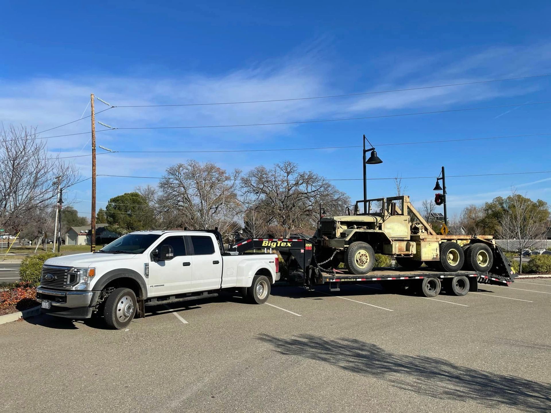White pickup towing a military vehicle on a trailer in a parking lot under a blue sky