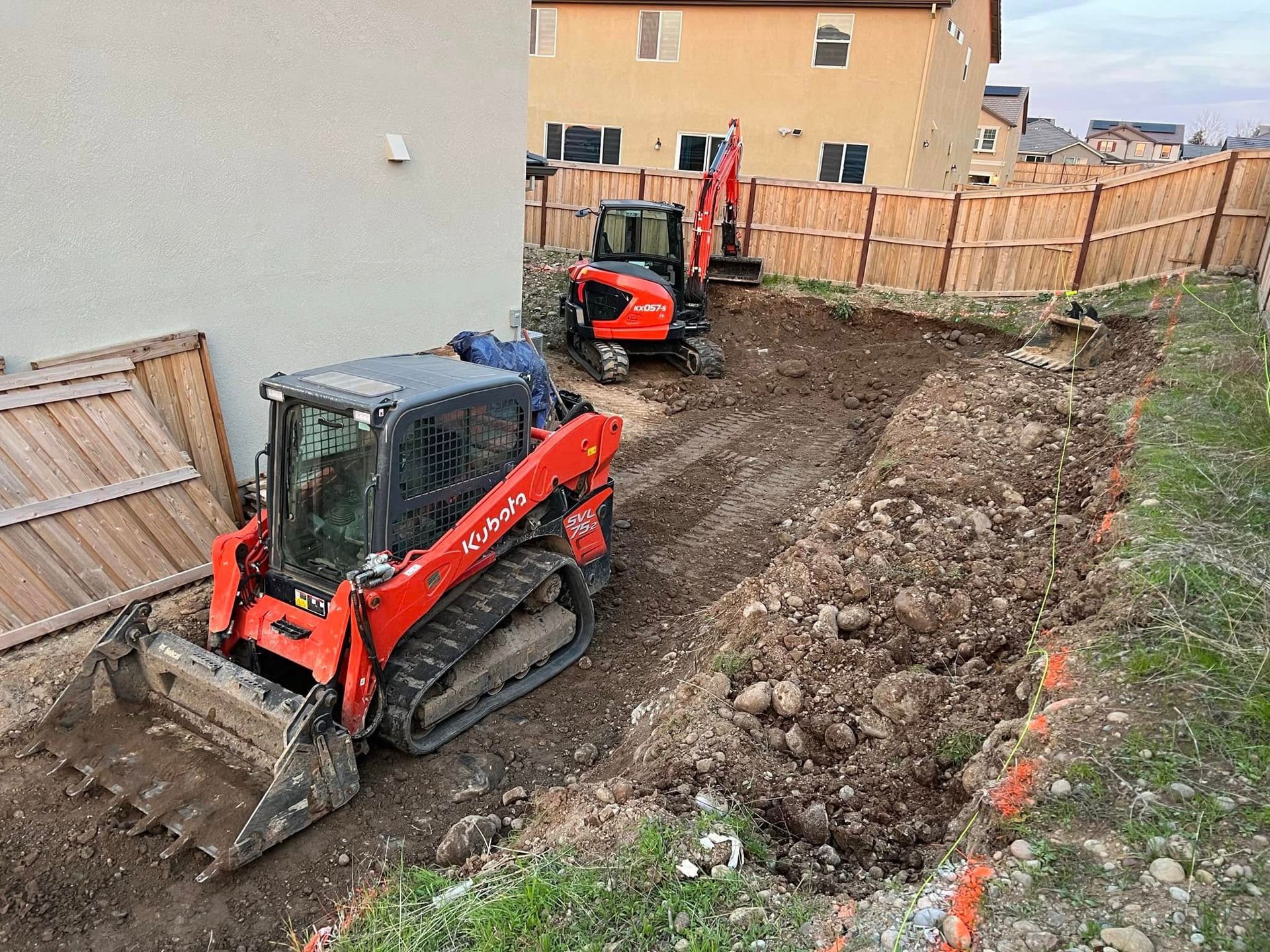Two red mini excavators working in a dirt backyard beside a fence and house.