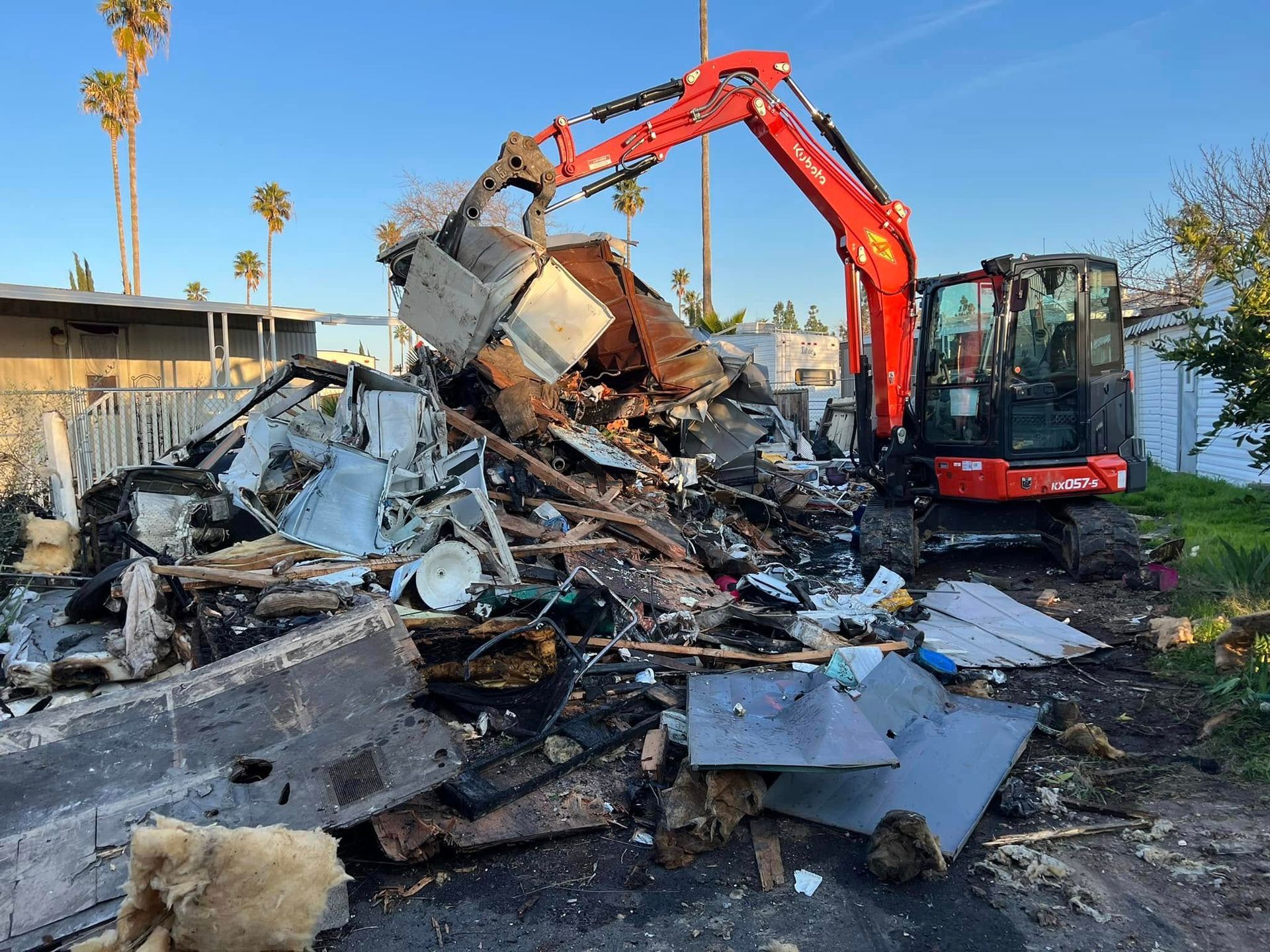 Excavator demolishing a burned-down structure, with rubble piled high under a clear blue sky.