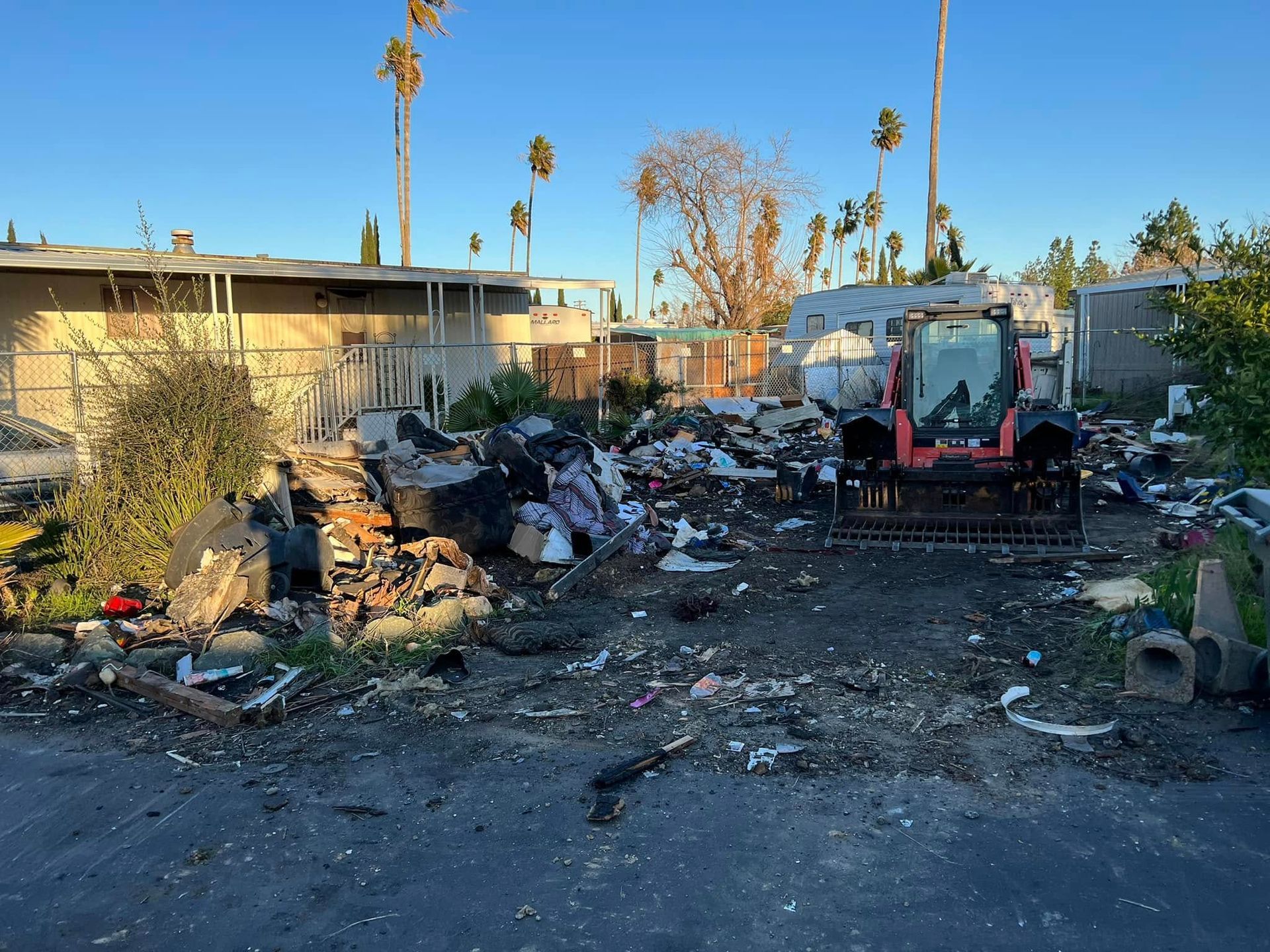 Trash pile and debris in a yard beside a small excavator, with houses and palm trees in the background.