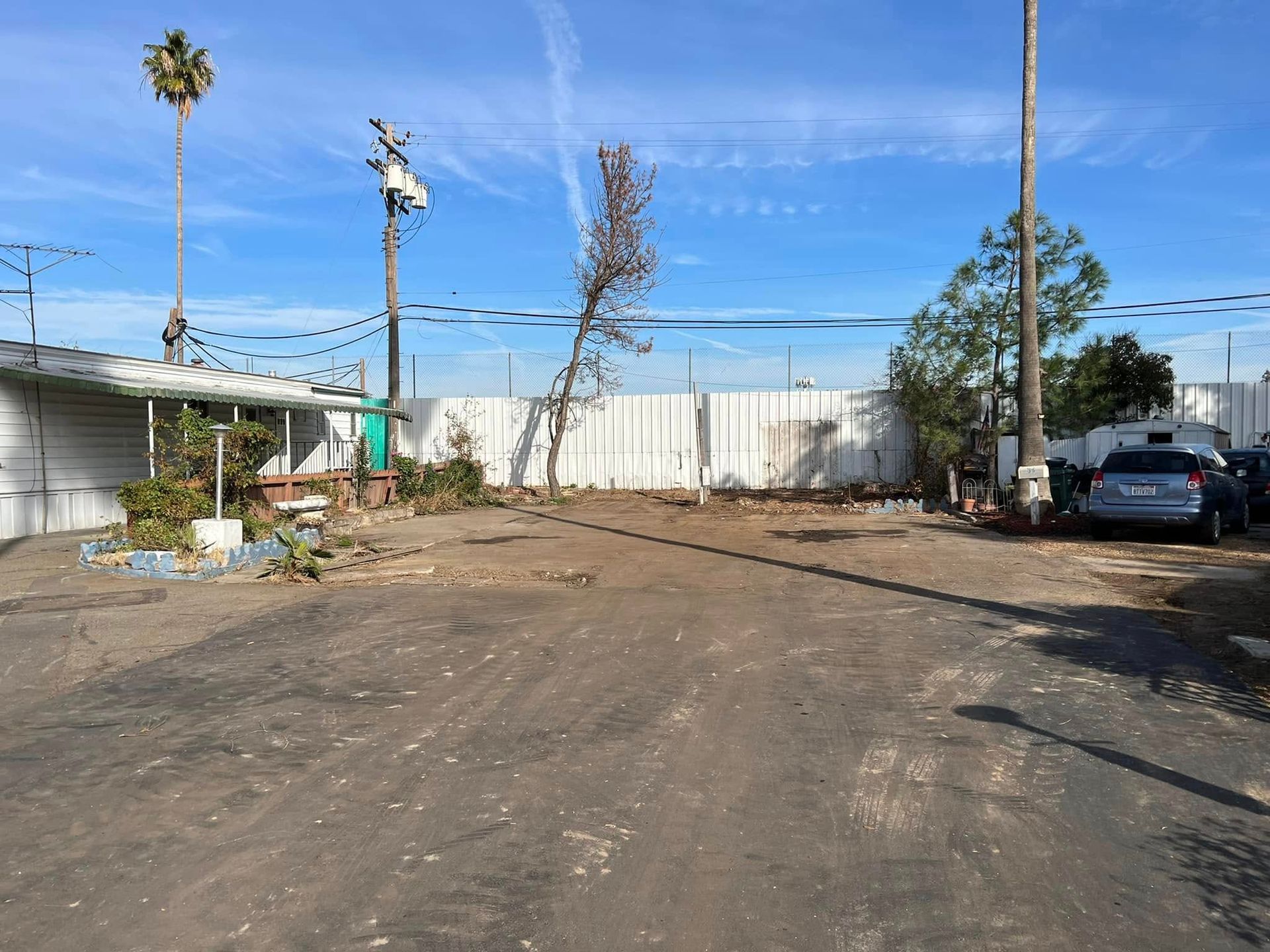 Dusty vacant lot with parked cars, palm trees, and a white fence under a blue sky