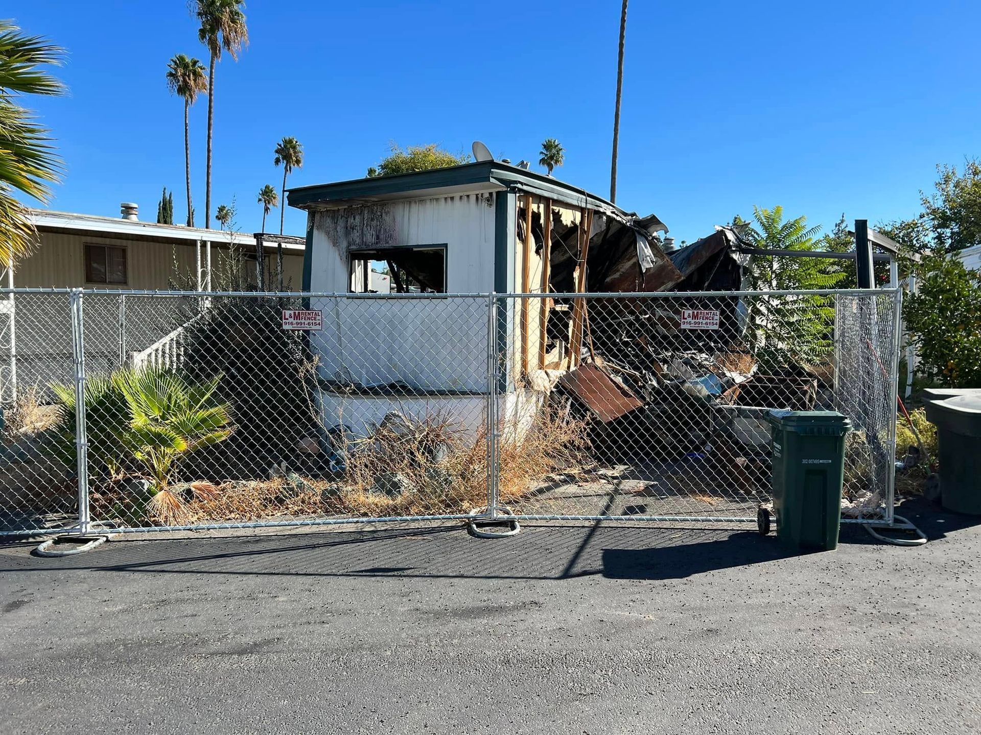 Abandoned house with burned-out roof behind a chain-link fence on a sunny street