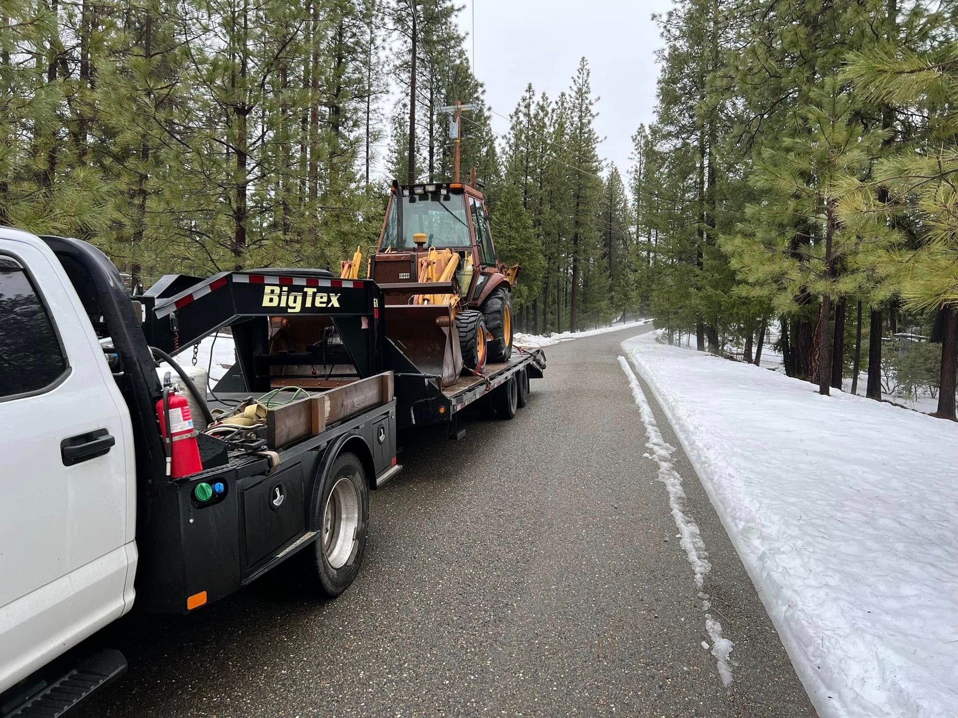 Tow truck hauling a car on a snowy forest road beside a snowbank and pine trees
