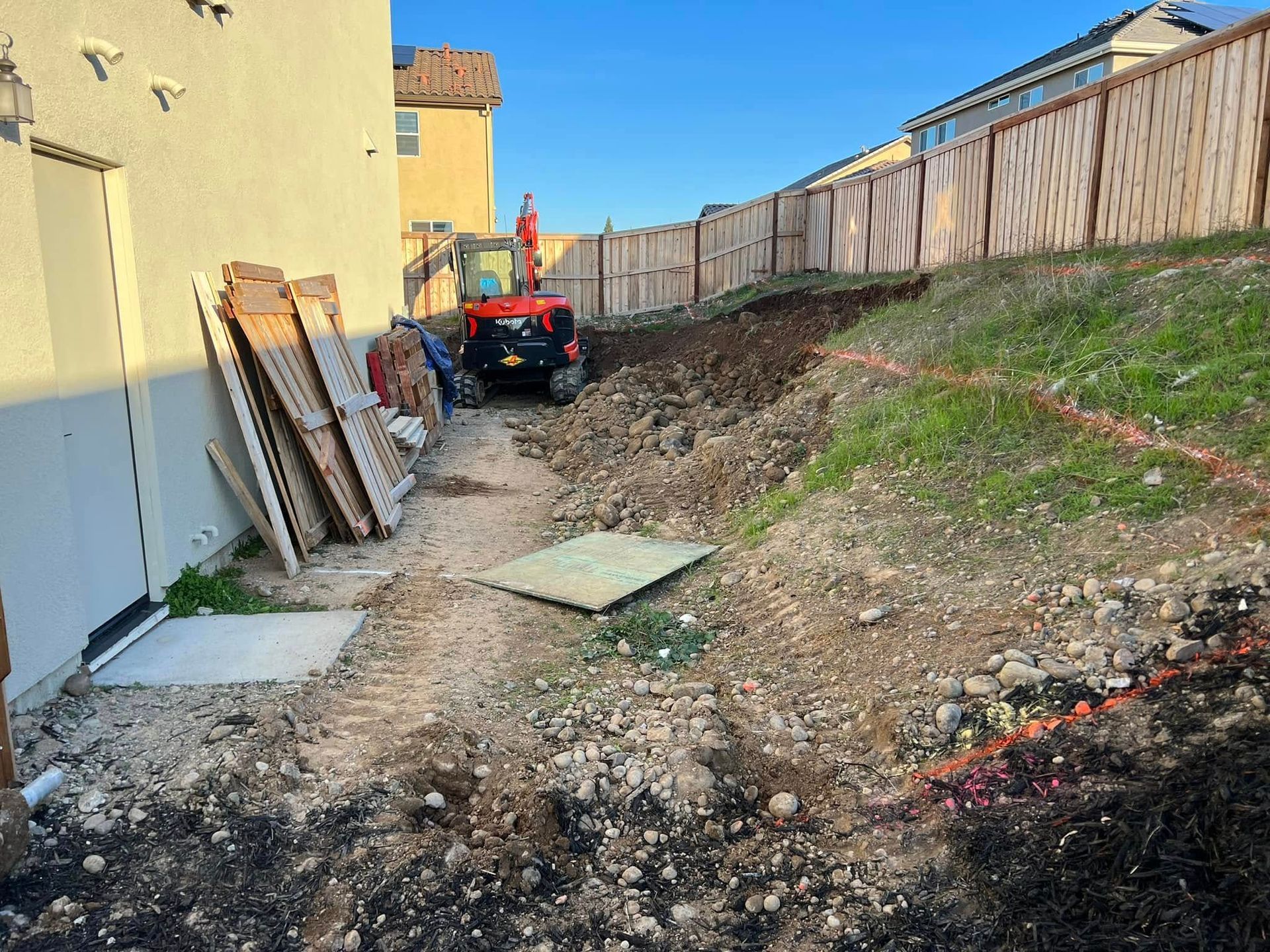 Narrow dirt path between a house and fence, with construction materials and a small tractor at the end.