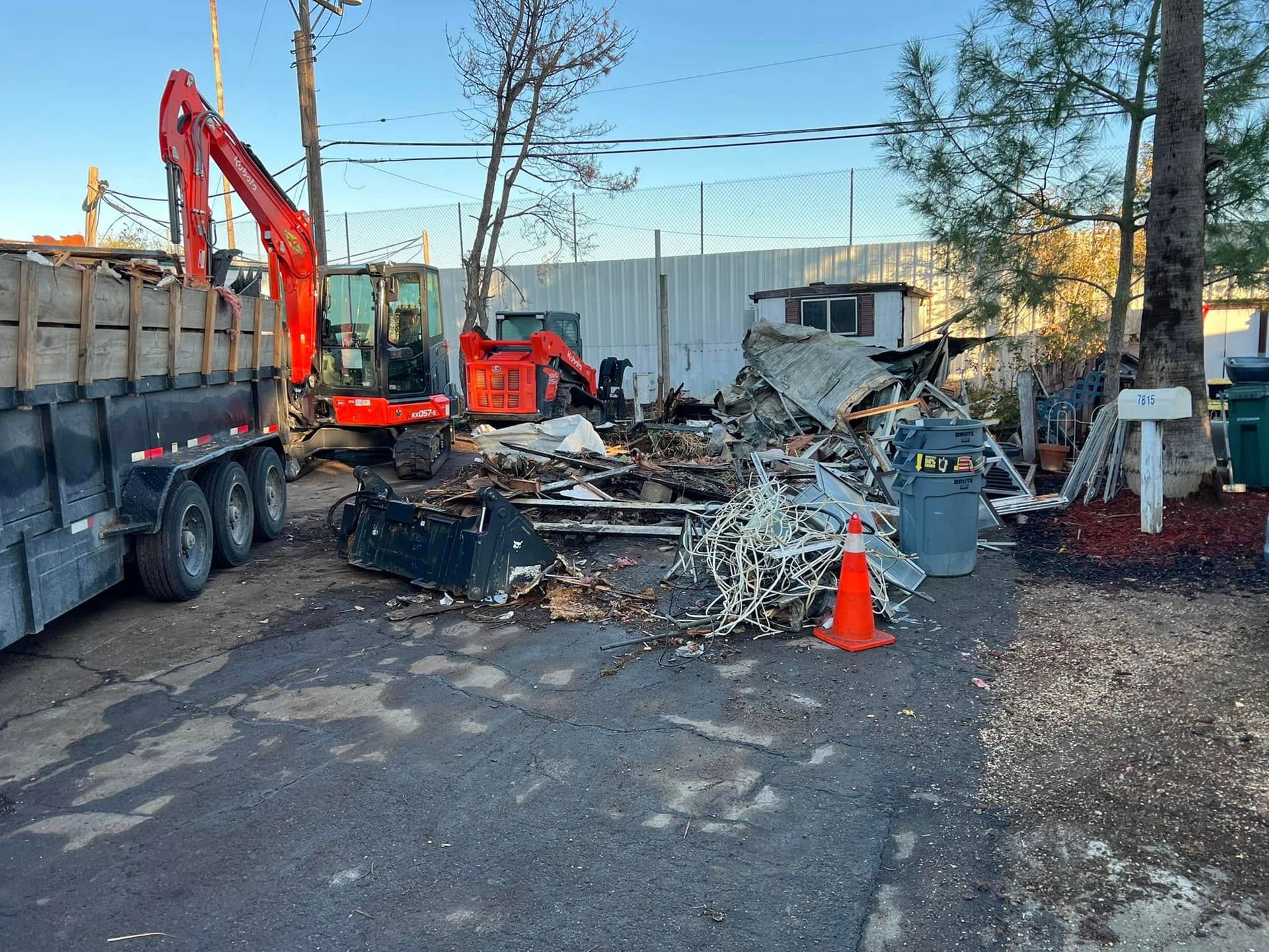 Demolition debris with red excavator, dump truck, and orange cone beside a damaged building site
