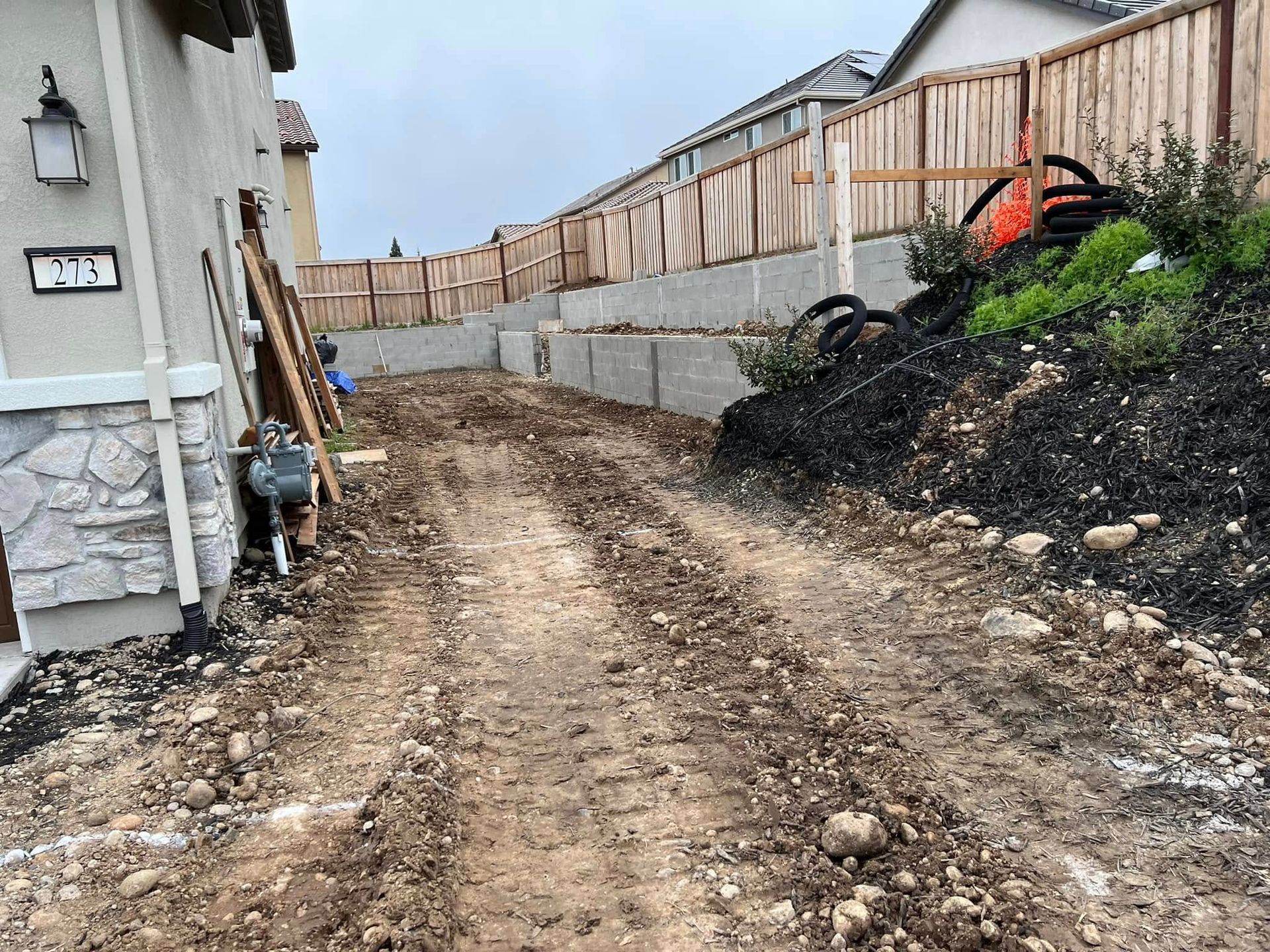 Dirt driveway under construction beside a house, with retaining wall, fencing, and utility pipes.