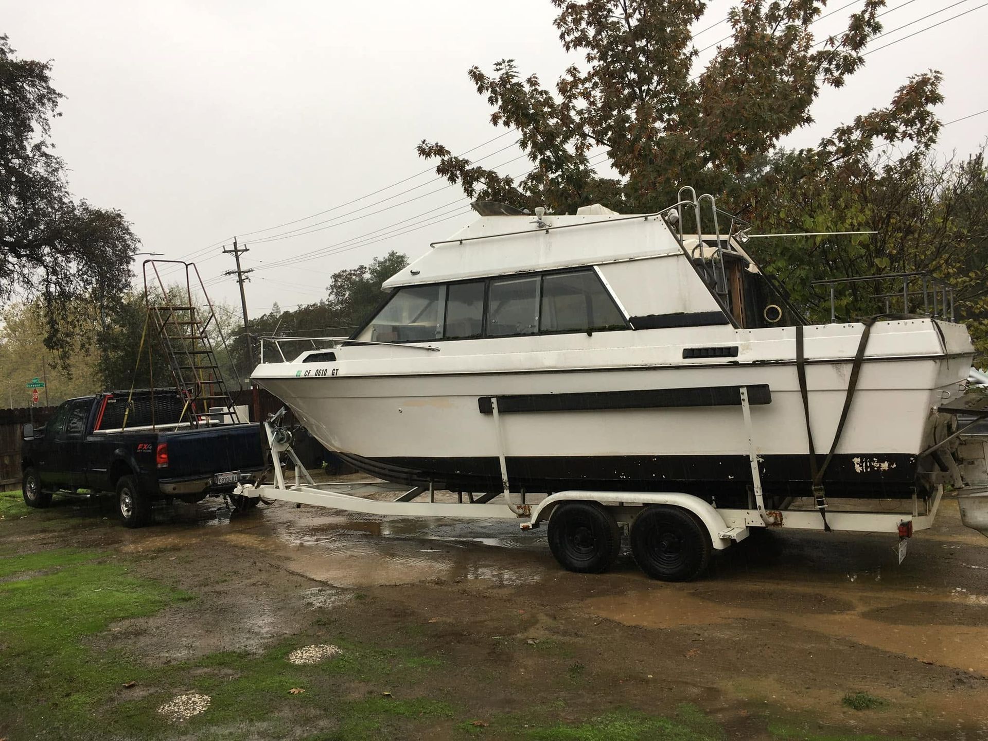 White cabin cruiser on a trailer in a muddy yard, with a black pickup truck nearby under overcast skies