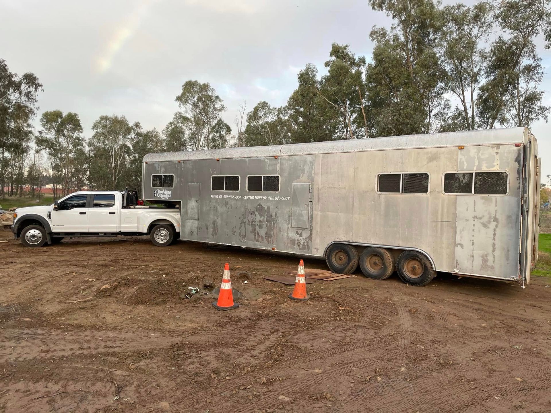 White pickup towing a long silver trailer on a muddy dirt lot, with orange cones and trees in the background