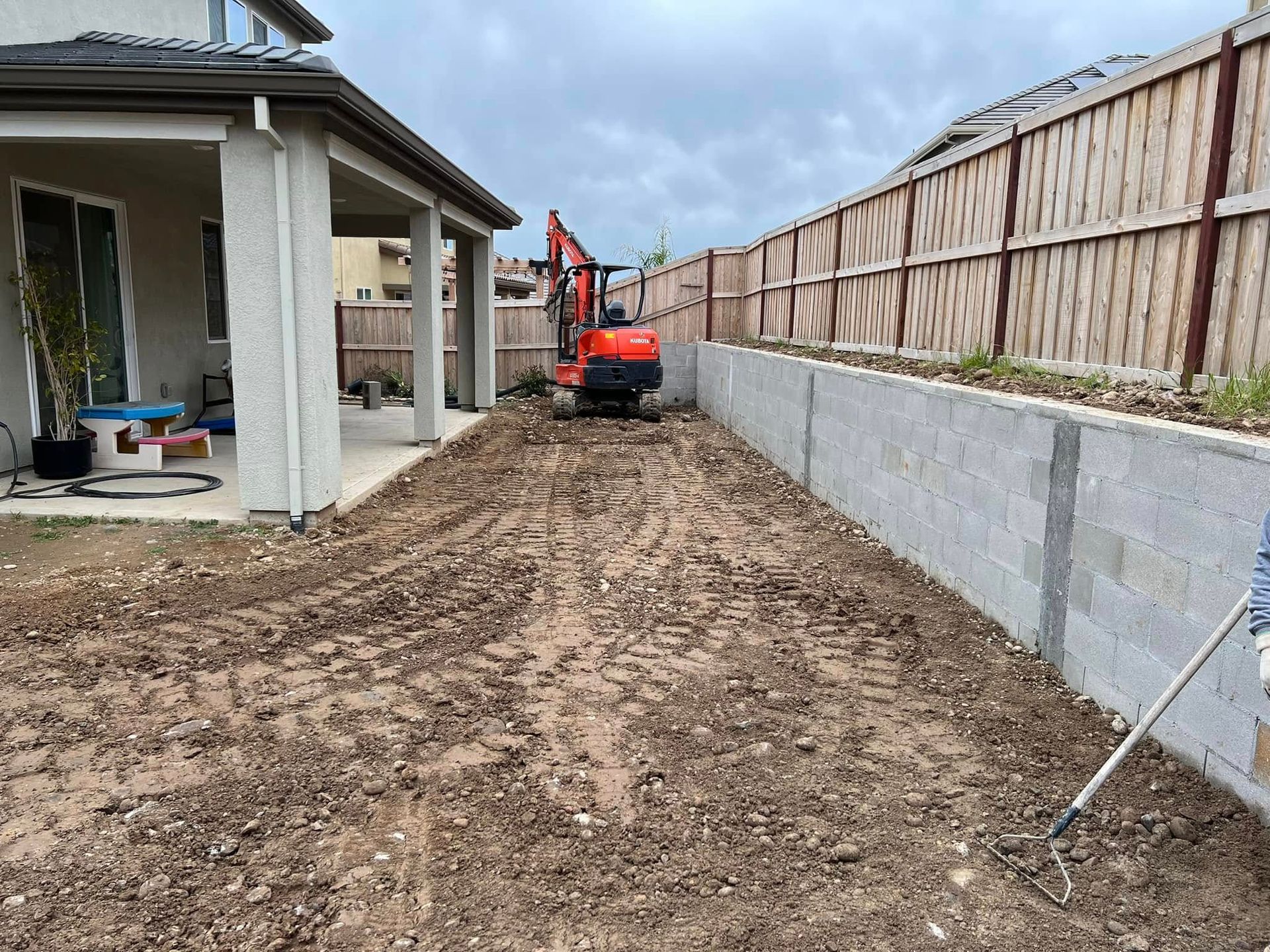 Backyard construction site with a mini excavator grading dirt beside a house and fence