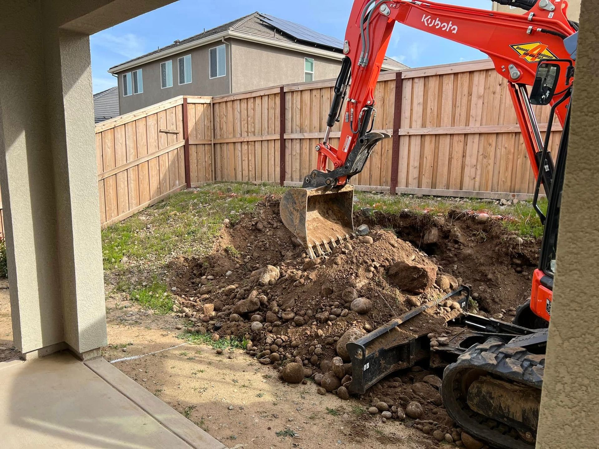 Orange excavator digging a backyard hole beside a wooden fence and house patio area