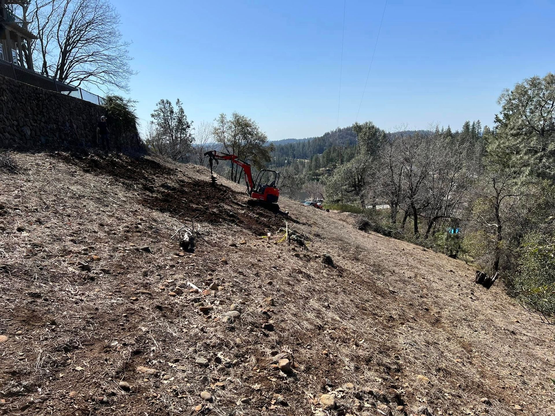 Dry hillside with a red excavator clearing brush under a clear blue sky