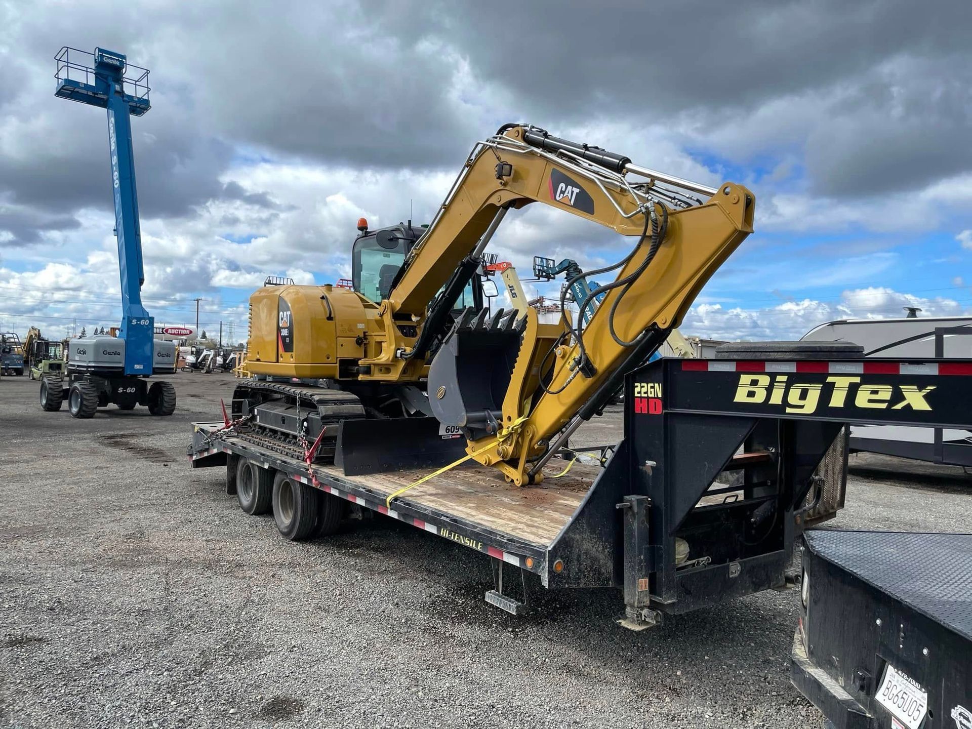 Yellow excavator loaded on a Big Tex trailer in a gravel lot under cloudy skies.