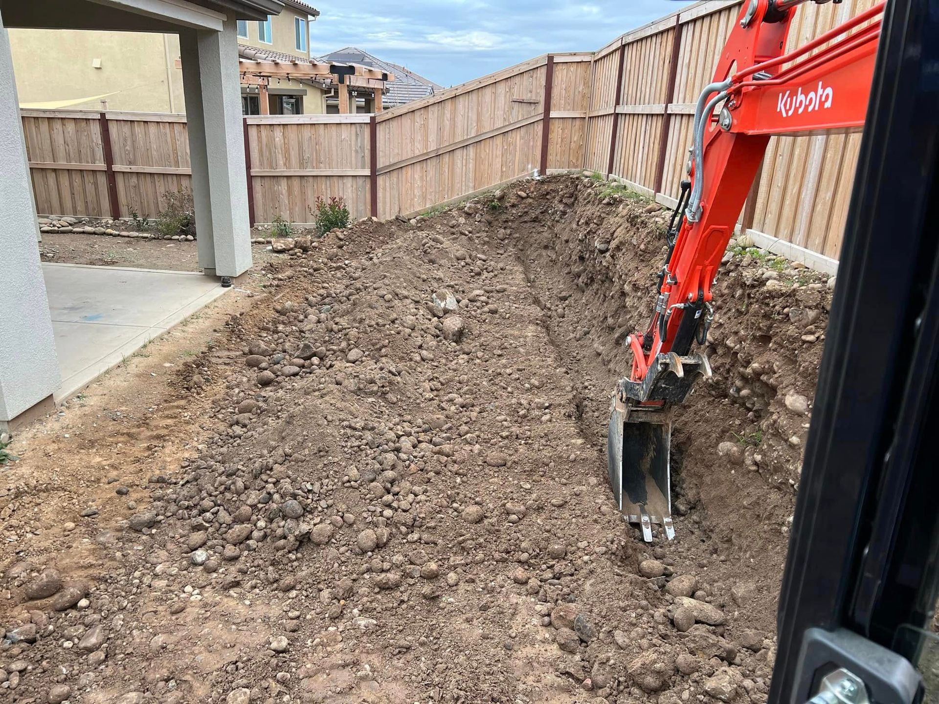 Backyard excavation with a red excavator digging a large dirt mound beside a wooden fence