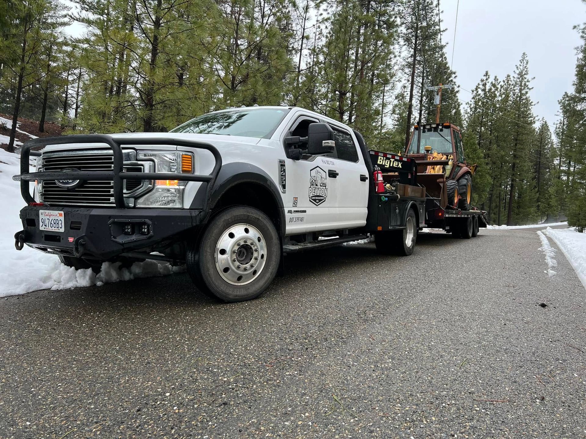 White tow truck hauling equipment trailer on a gravel forest road under overcast sky