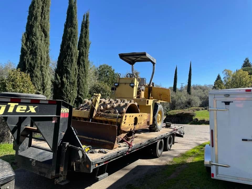 Yellow bulldozer on a flatbed trailer beside a white truck on a sunny roadside with tall cypress trees