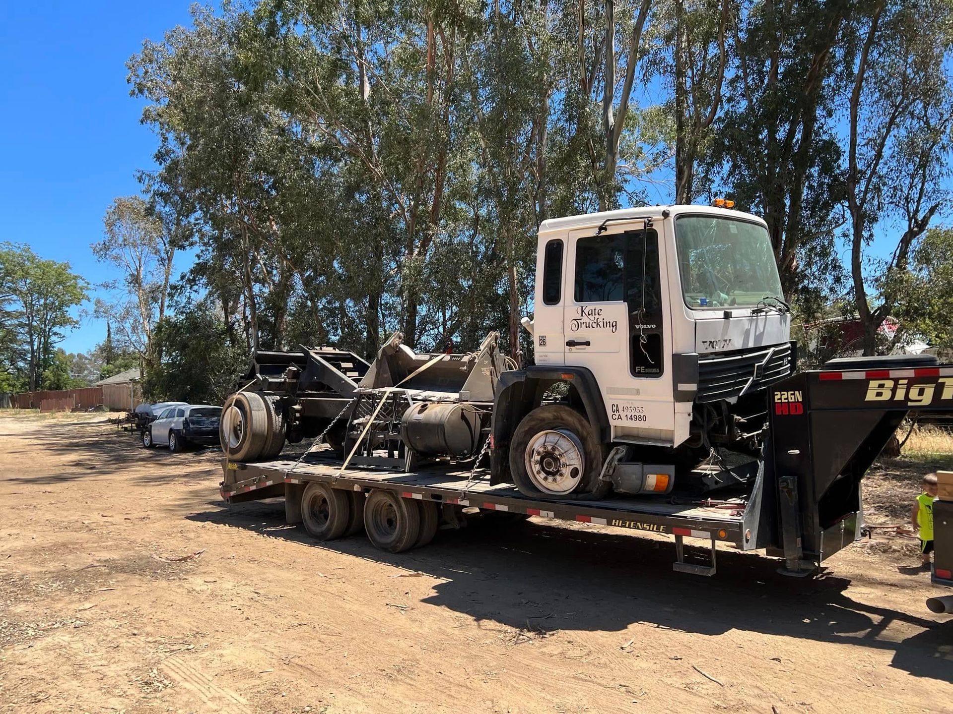 Truck chassis loaded on a flatbed trailer in a dirt lot under trees.