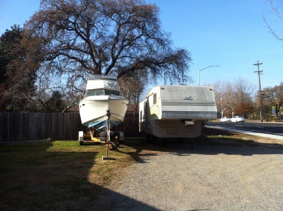Boat and RV parked on a gravel driveway beside a wooden fence and bare trees