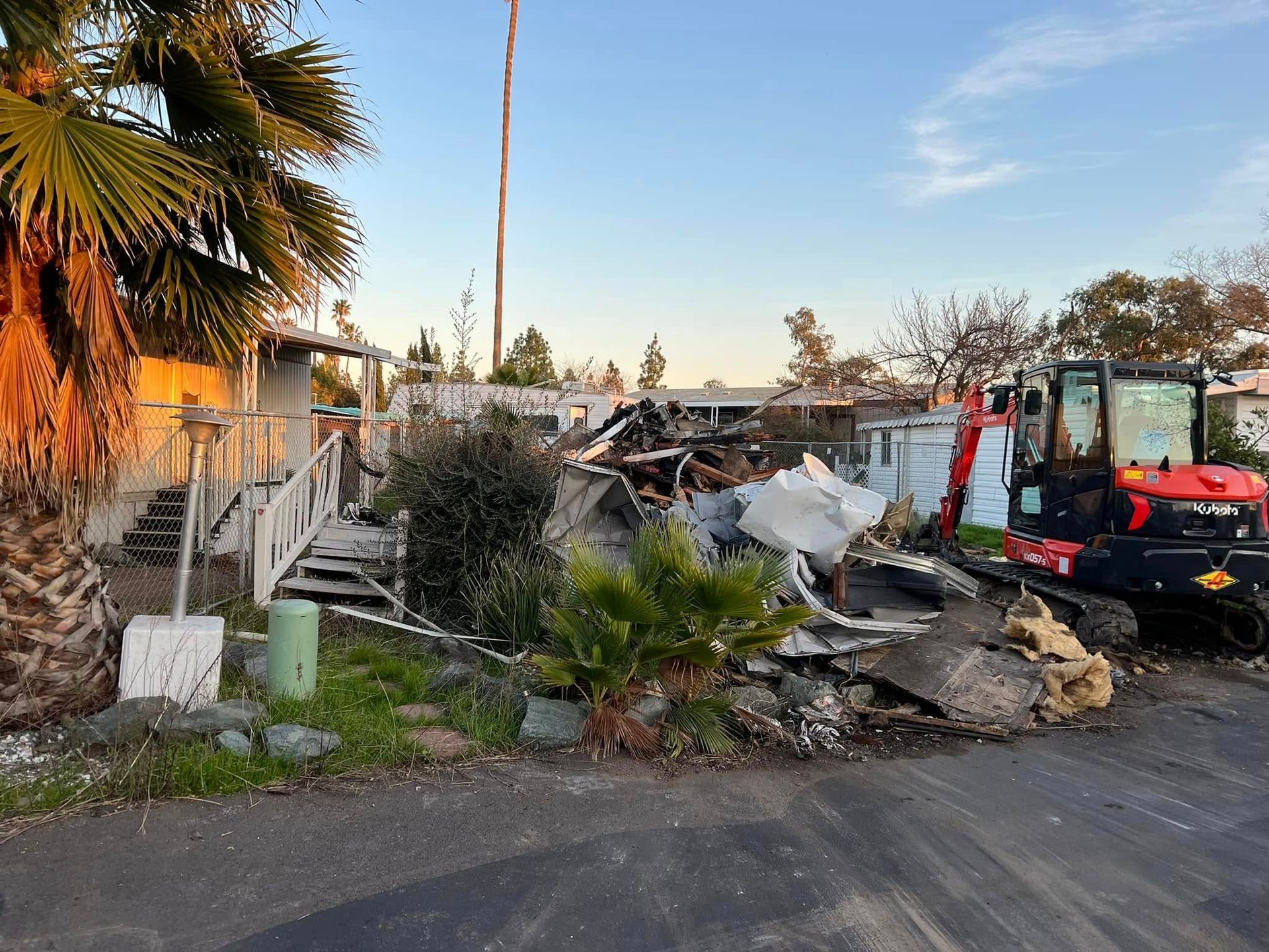 Rubble and debris in a yard beside a road, with a small red excavator and palm trees at sunset