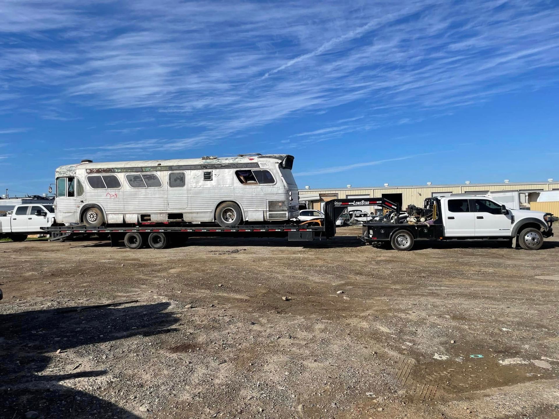 White RV on a flatbed tow truck with a white pickup in a gravel lot under a blue sky