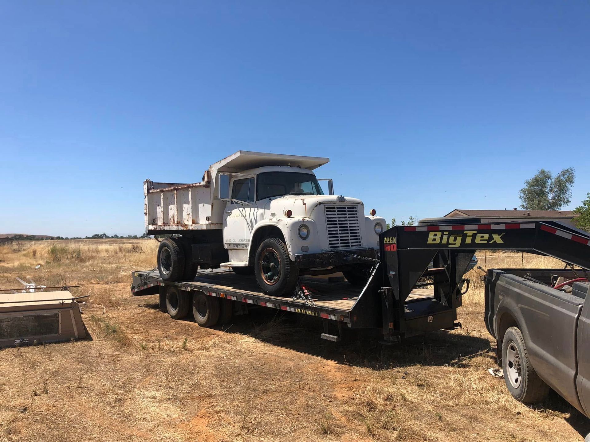 White dump truck on a Big Tex trailer being towed across a dry field under a clear blue sky.