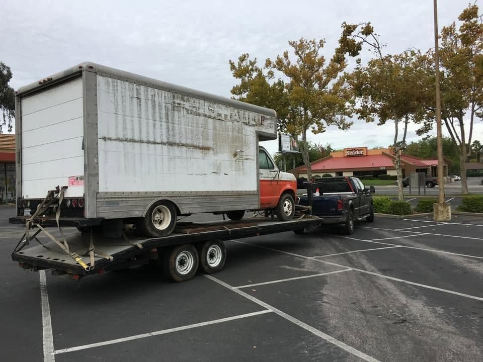 White box truck loaded on a flatbed trailer in a parking lot, with a black SUV nearby.