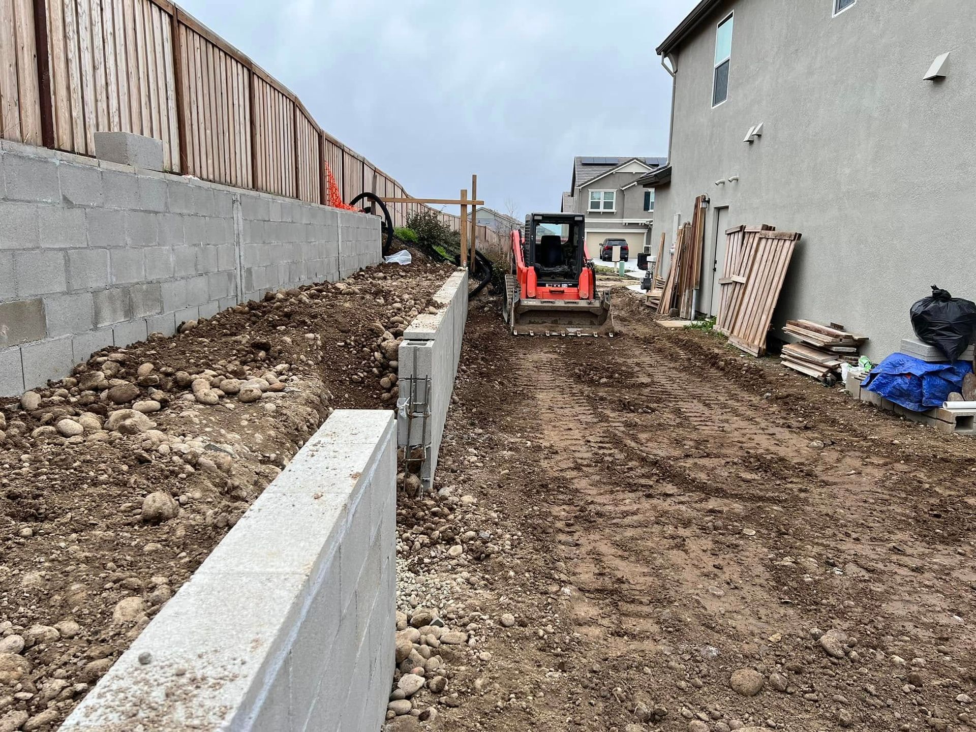 Narrow dirt construction path beside concrete walls, with a red skid steer ahead and building materials on the right.