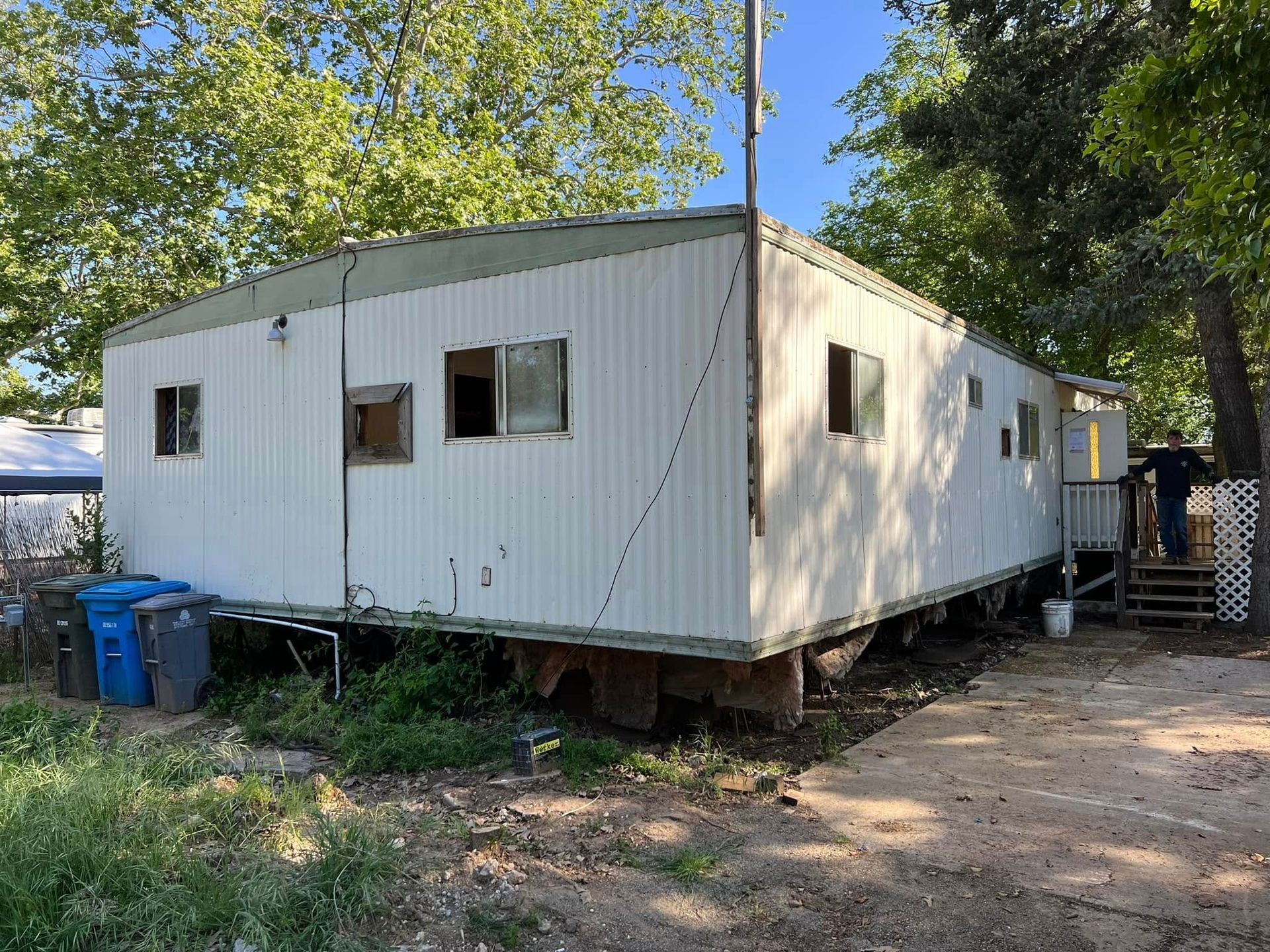 White portable building on raised blocks in a wooded yard with a dirt driveway and blue bins