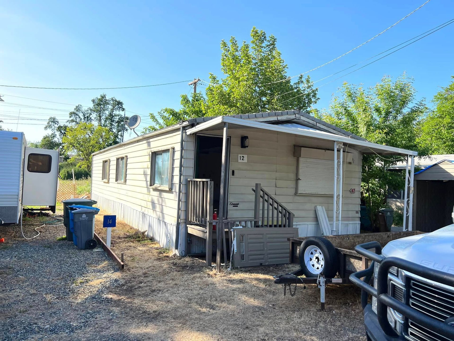 Small beige mobile home with a porch and ramp in a gravel lot, with trees and a truck nearby