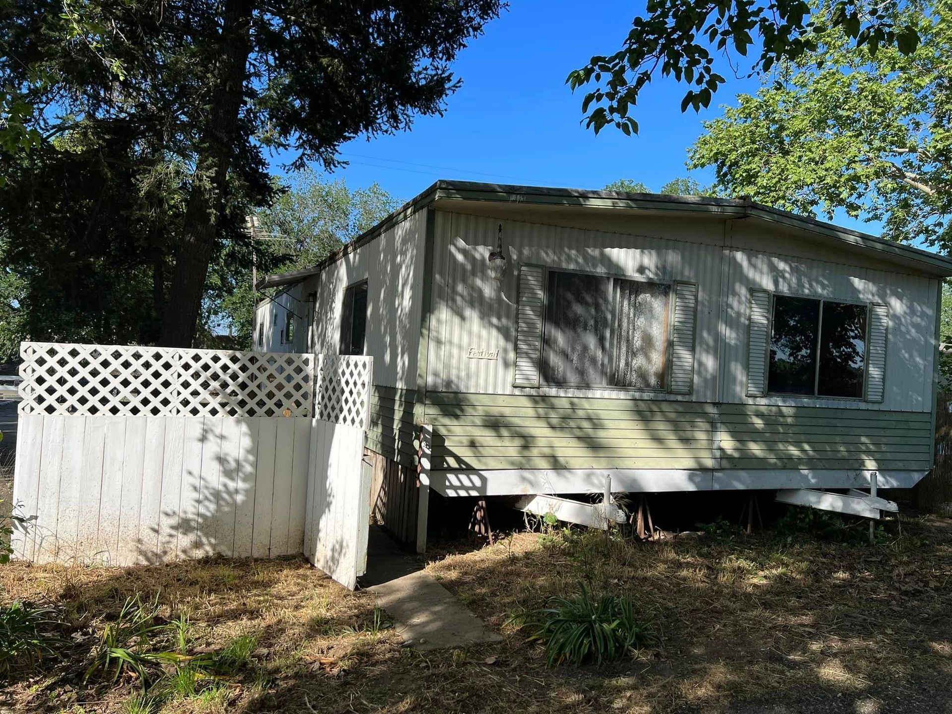 Small light-green house with a white fence and lattice, shaded by trees in a yard.