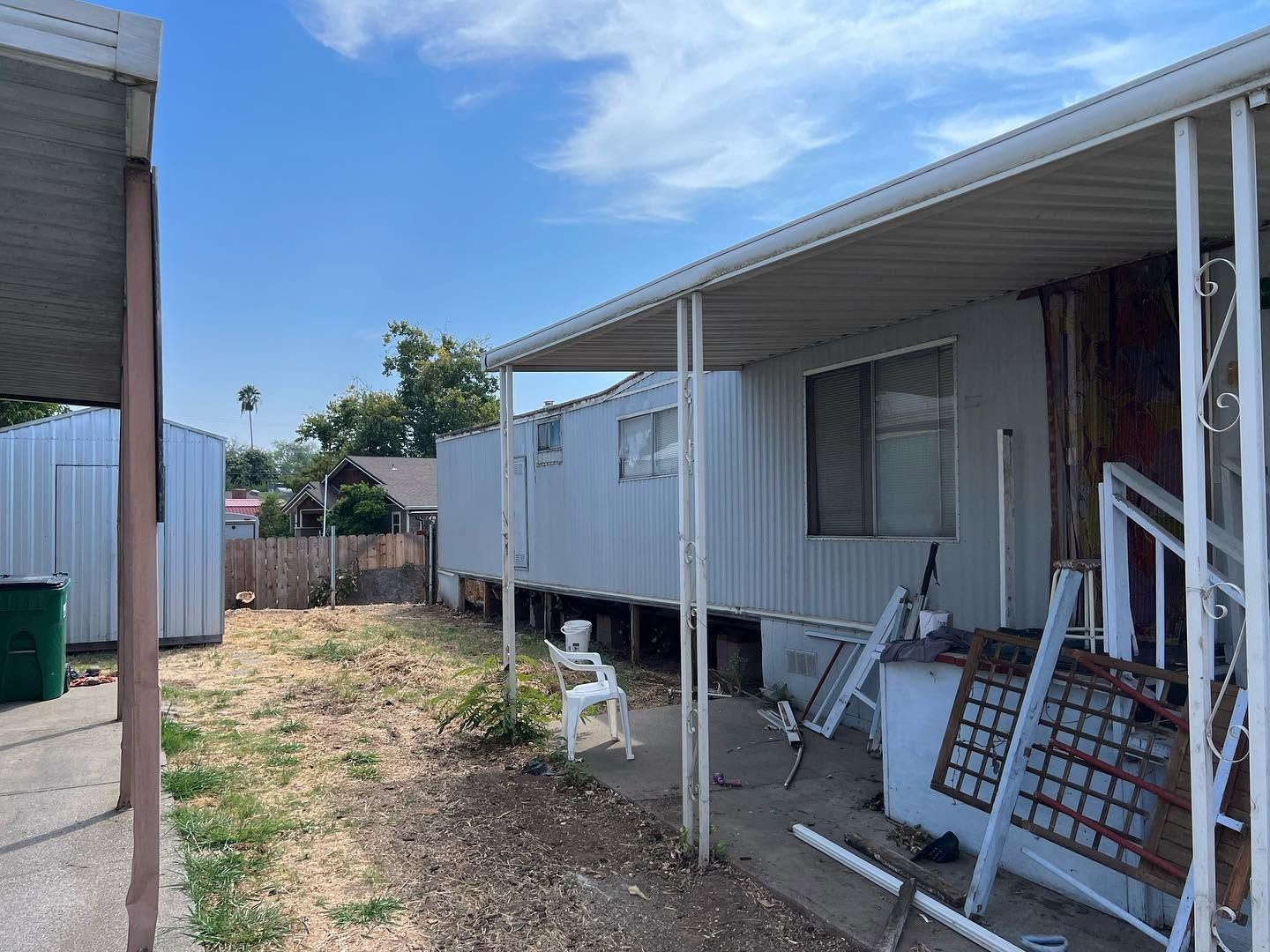 Mobile home with a covered porch and scattered household items in a dirt yard, under a blue sky