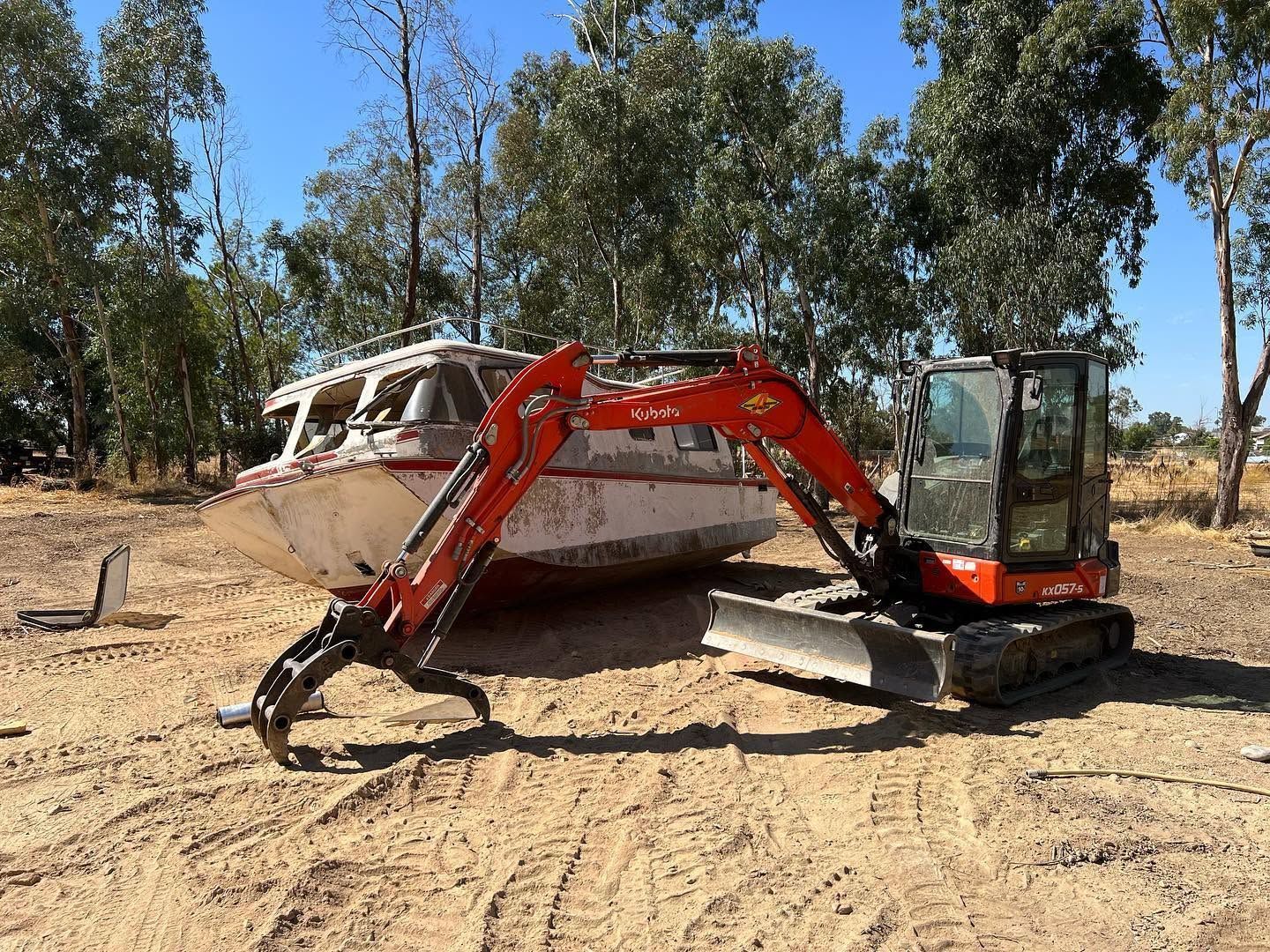 Red excavator beside a white boat on dry dirt in a wooded area