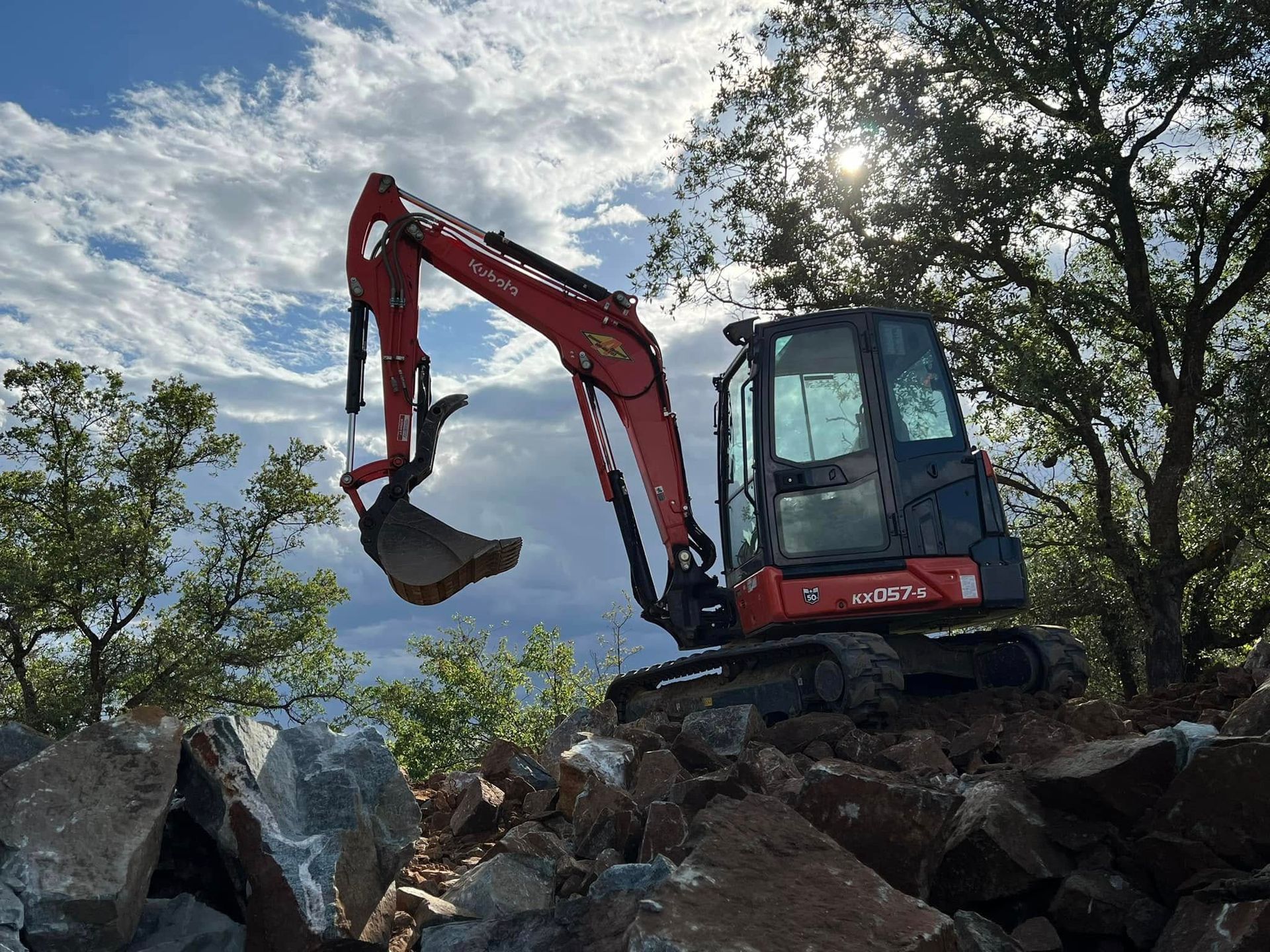 Red excavator perched on rocks lifting a bucket against a bright blue sky and trees.