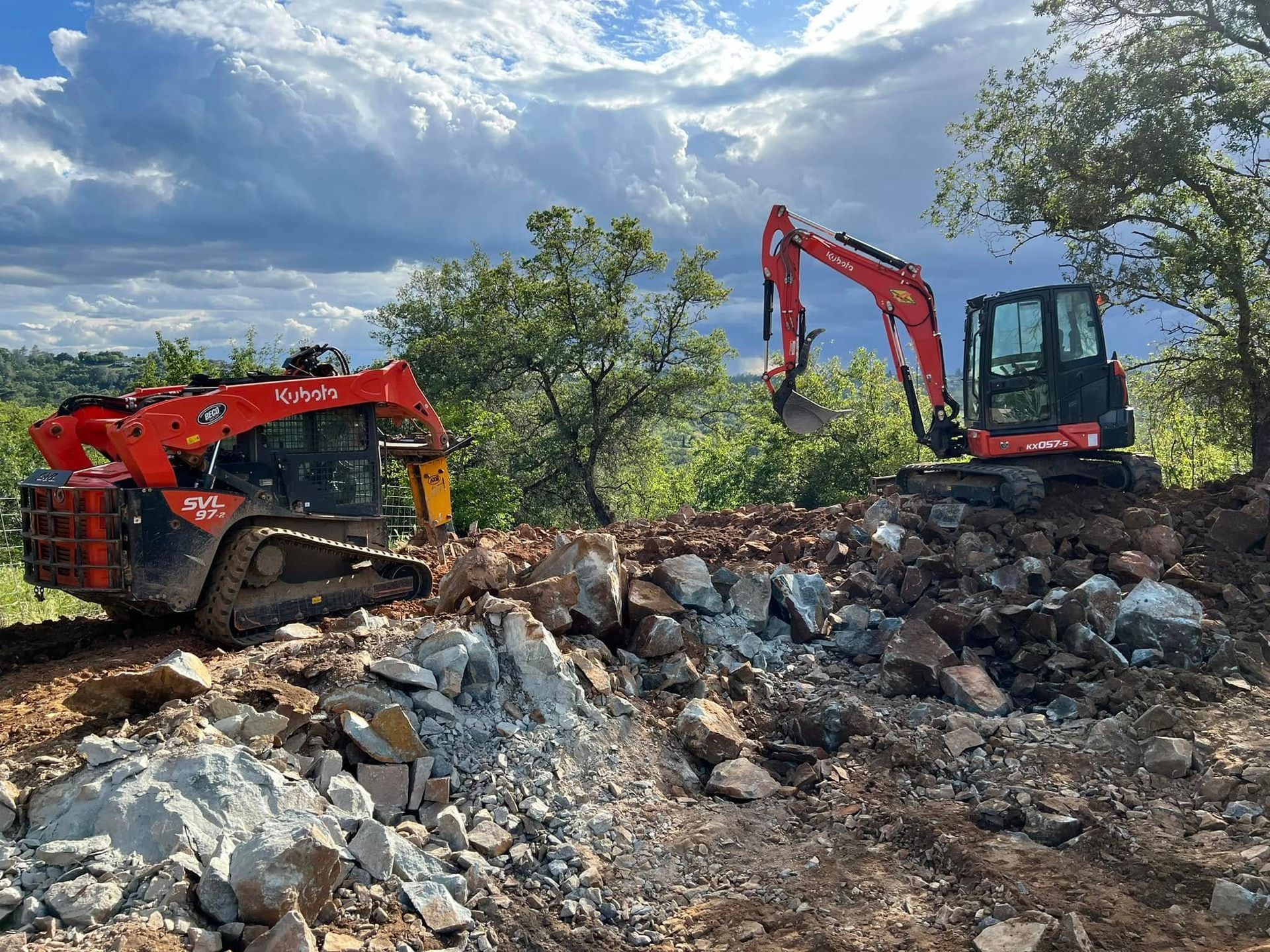 Two red excavators working on a rocky dirt embankment under a cloudy sky