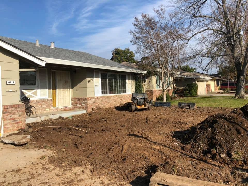 Front yard under renovation beside a brick house, with piles of dirt and a small tractor.