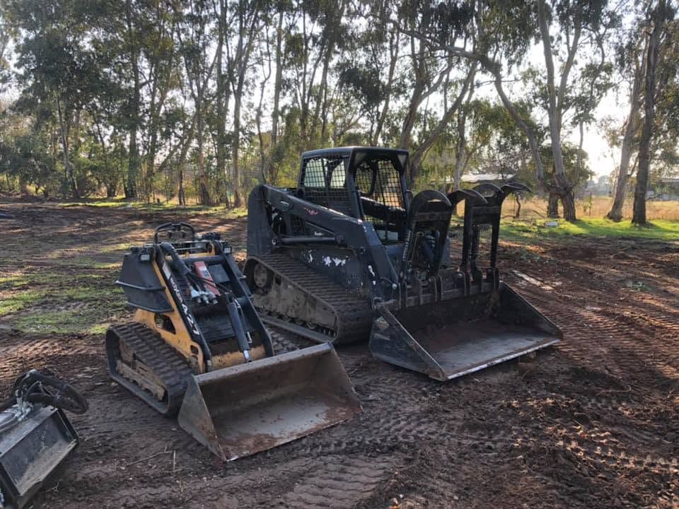 Two black tracked skid steer loaders with buckets parked on dirt in a wooded area