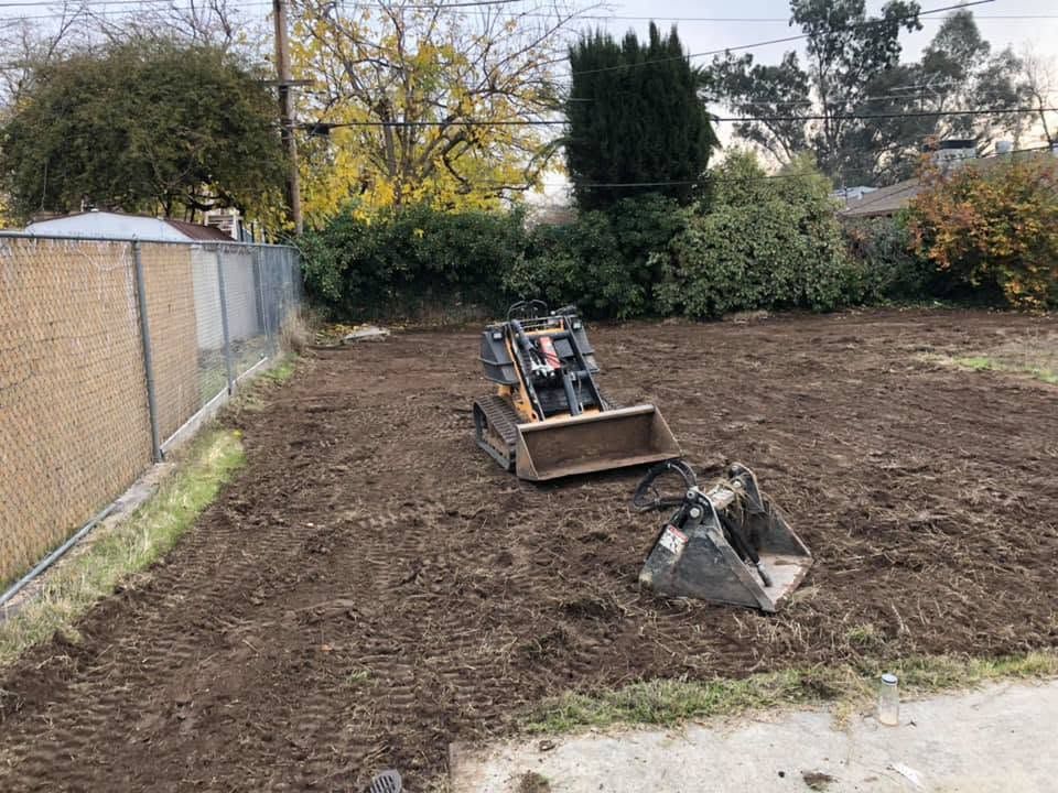 Construction equipment in a dirt backyard, with a loader bucket and a small pile of debris near a fence.