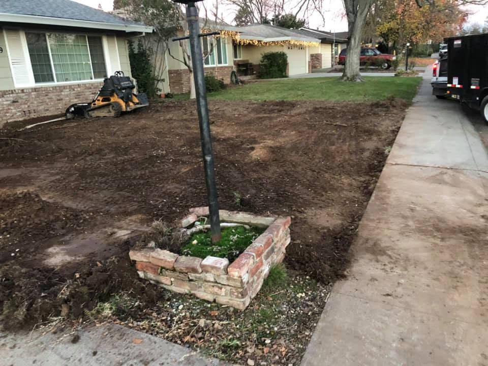 Front yard with a small brick planter, bare dirt, and a sidewalk beside a house and parked truck.