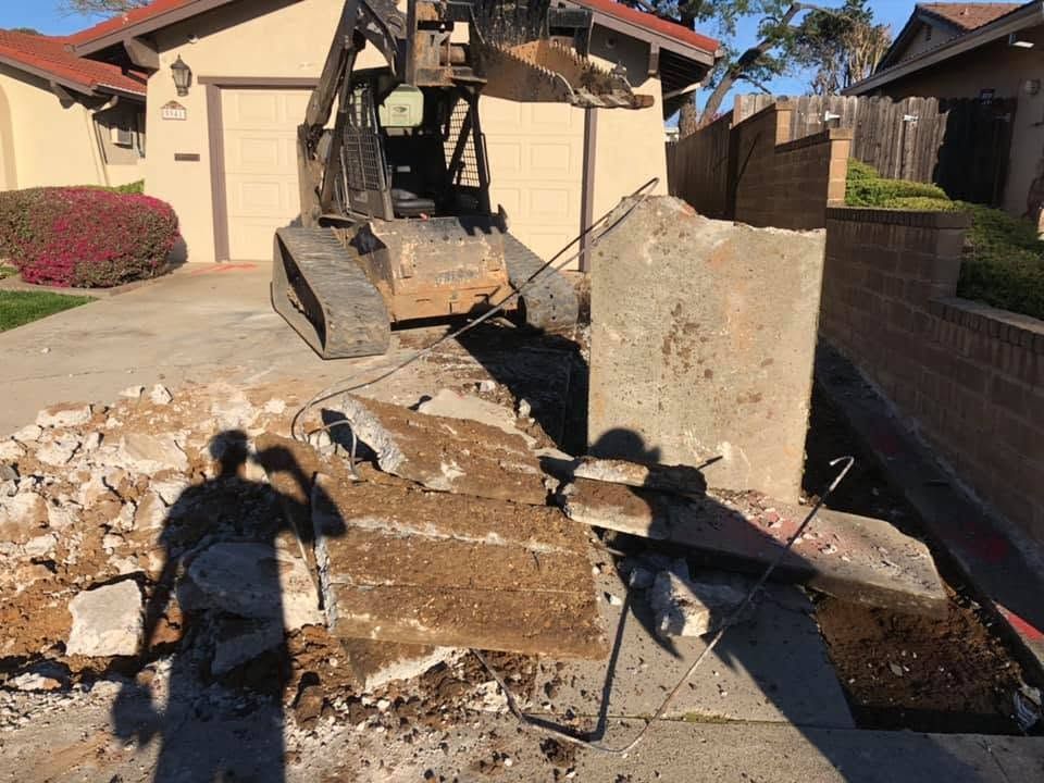 Construction skid steer removing concrete in a driveway beside a beige house and brick wall