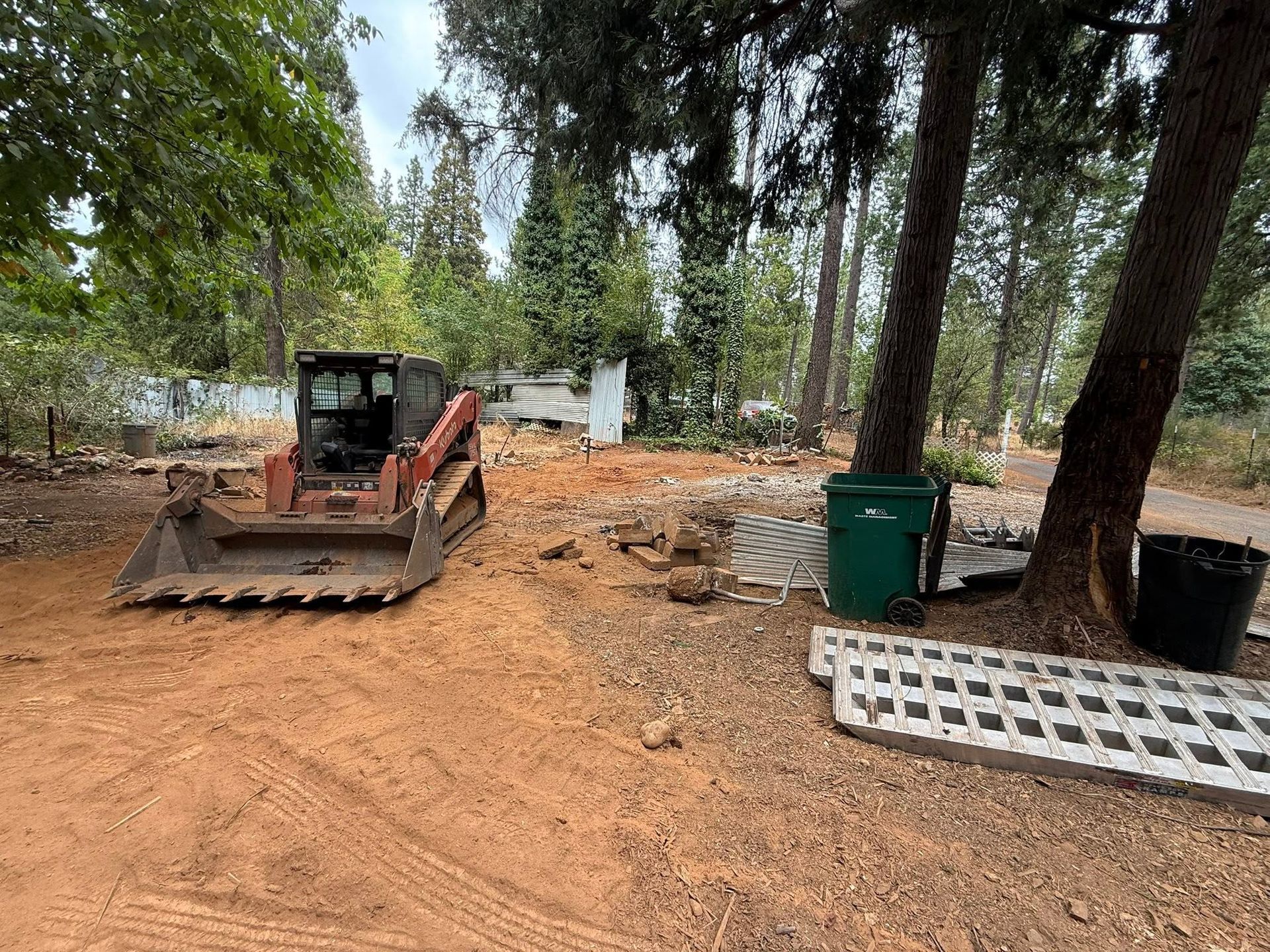 Bulldozer parked on a dirt lot among tall trees, with construction materials and a green bin nearby.