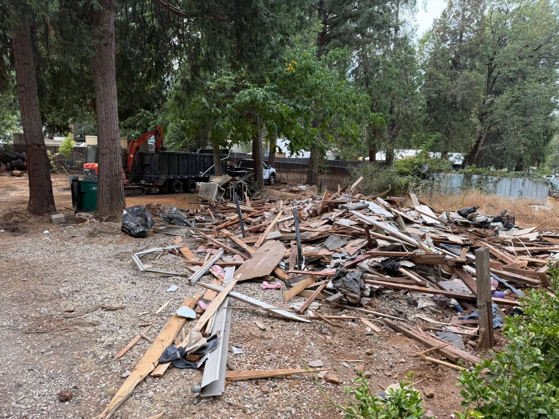 Pile of lumber and debris in a wooded yard with a trailer in the background