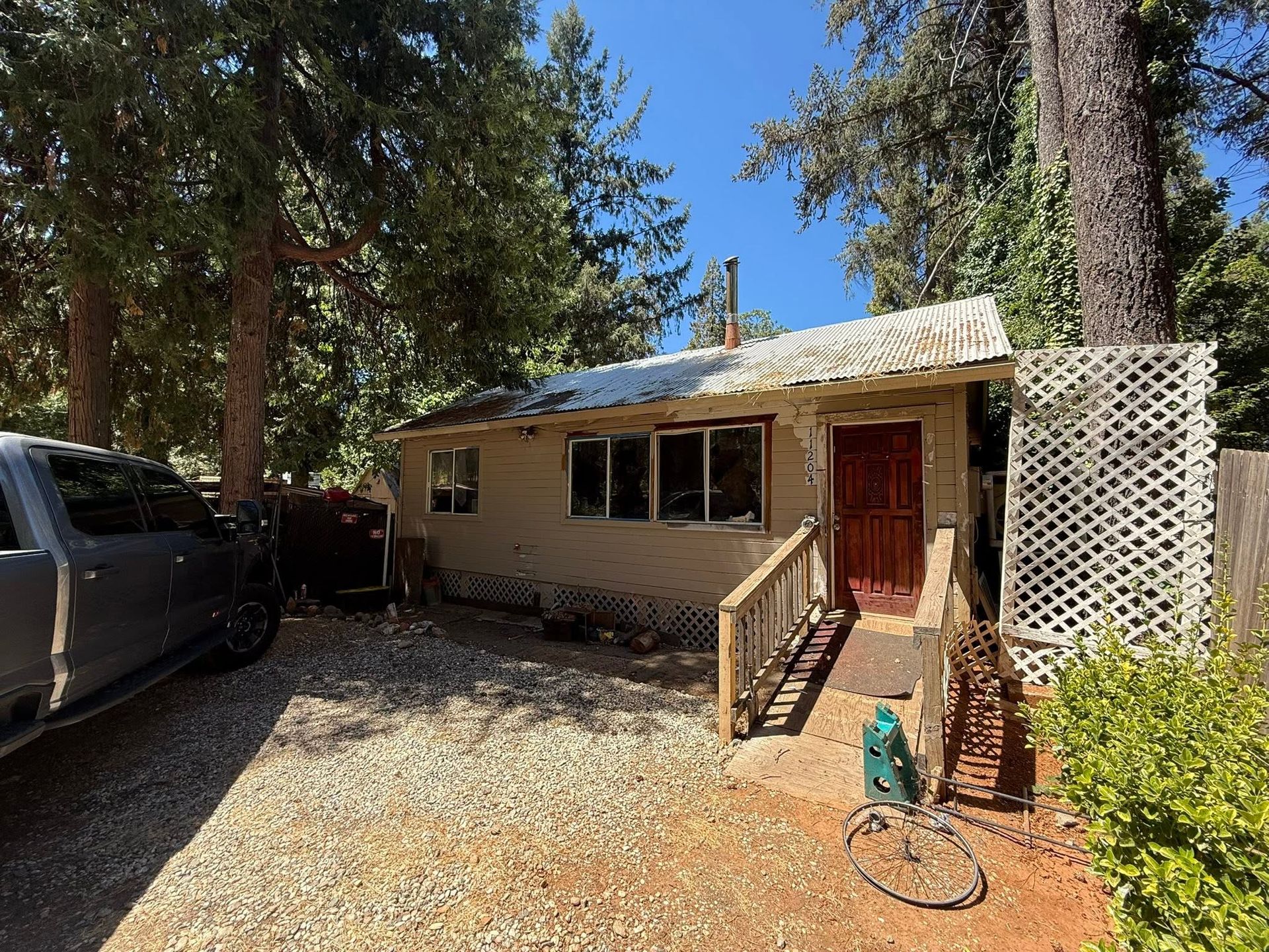 Small cabin with red door and lattice porch in a wooded area, with a gravel driveway and parked truck.